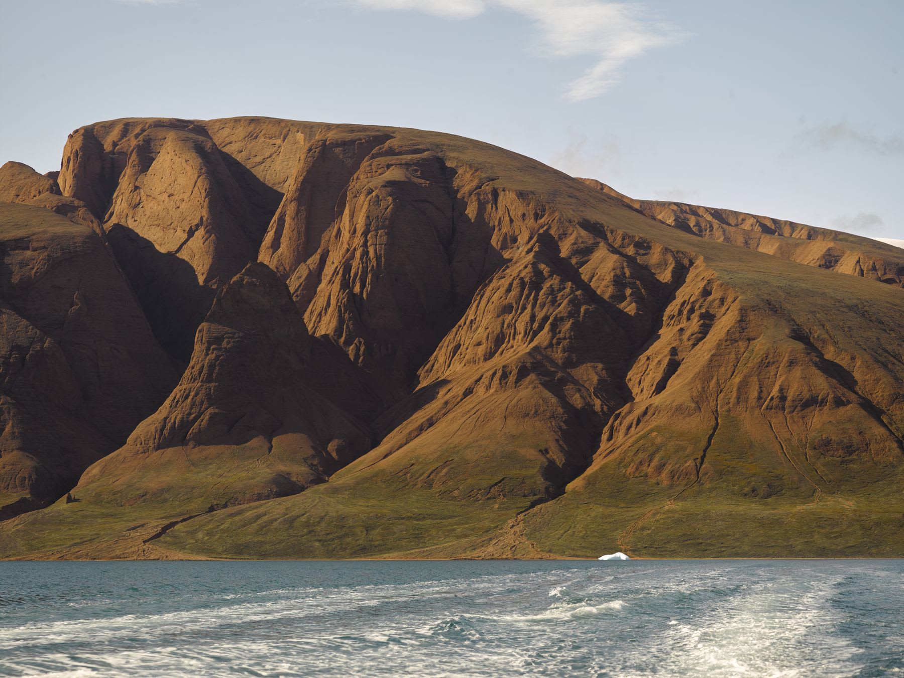 A vast mountain range rises steeply from the edge of a calm body of water, with the sun casting shadows across the rugged terrain. A faint trail of water behind a boat can be seen in the foreground under a clear sky.