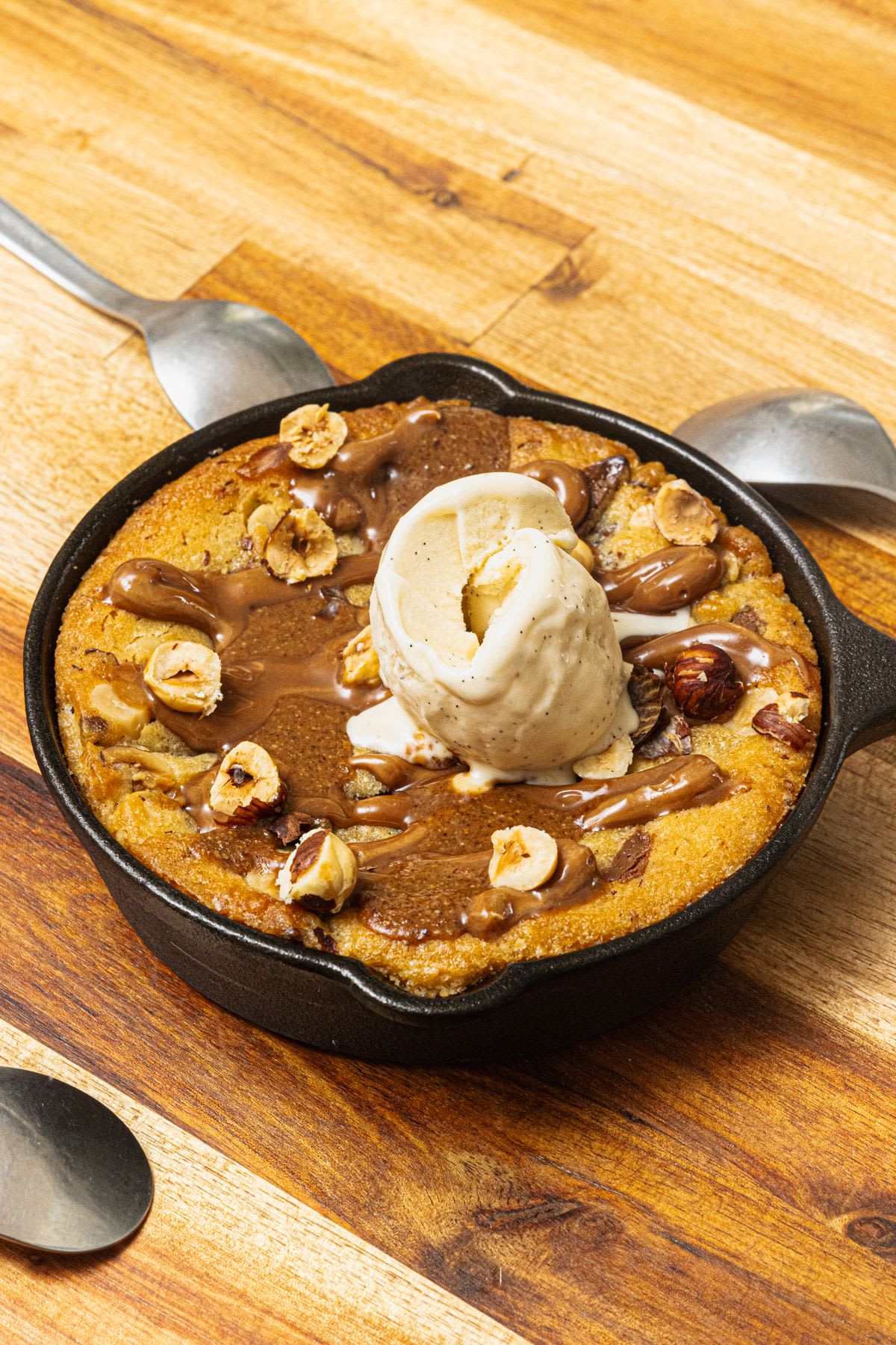 A skillet cookie topped with vanilla ice cream, chocolate or caramel sauce, and hazelnuts sits on a wooden surface, flanked by spoons.