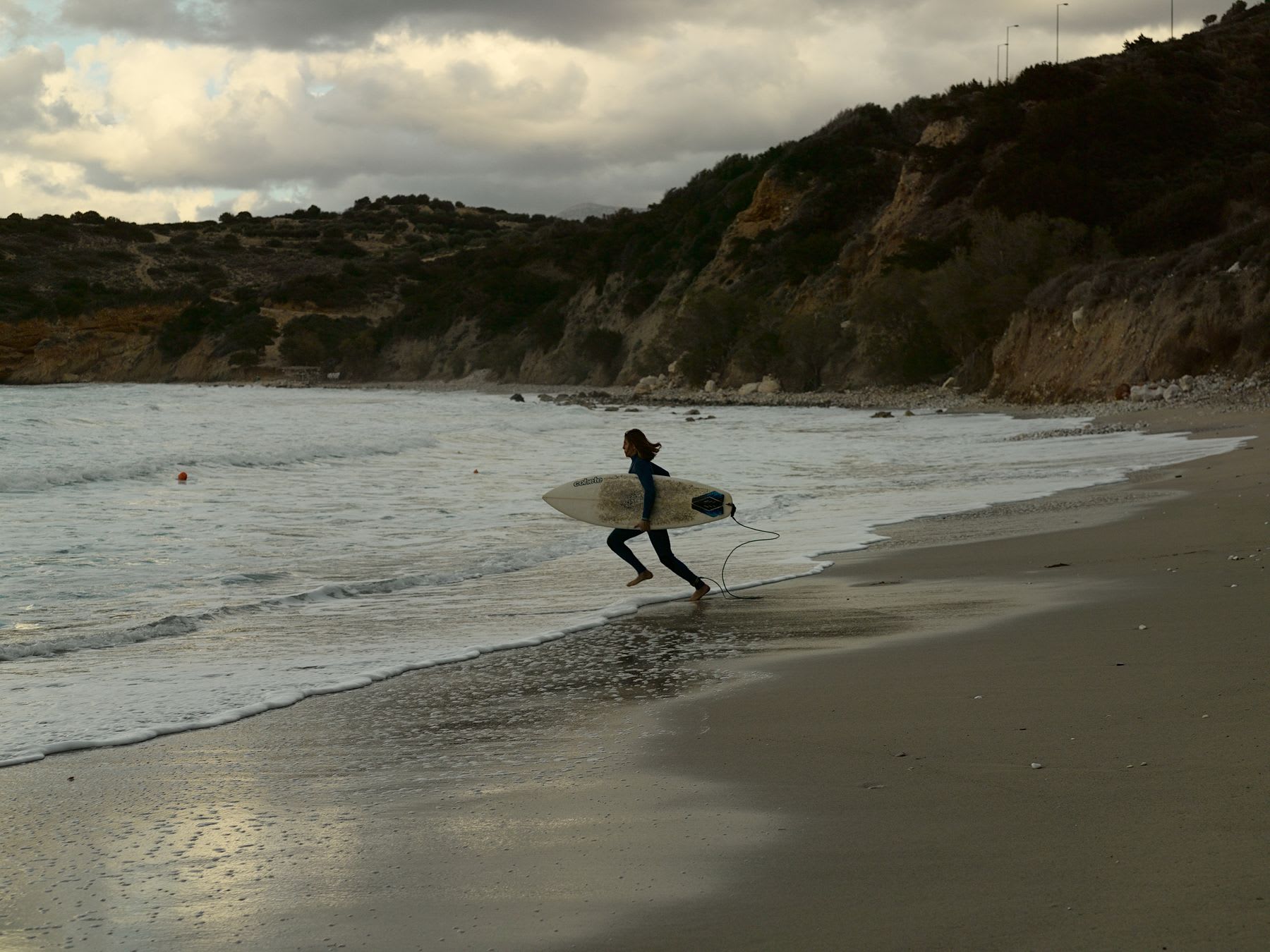 A surfer in a wetsuit runs along the shoreline with a surfboard as waves crash on the sandy beach under a cloudy sky.