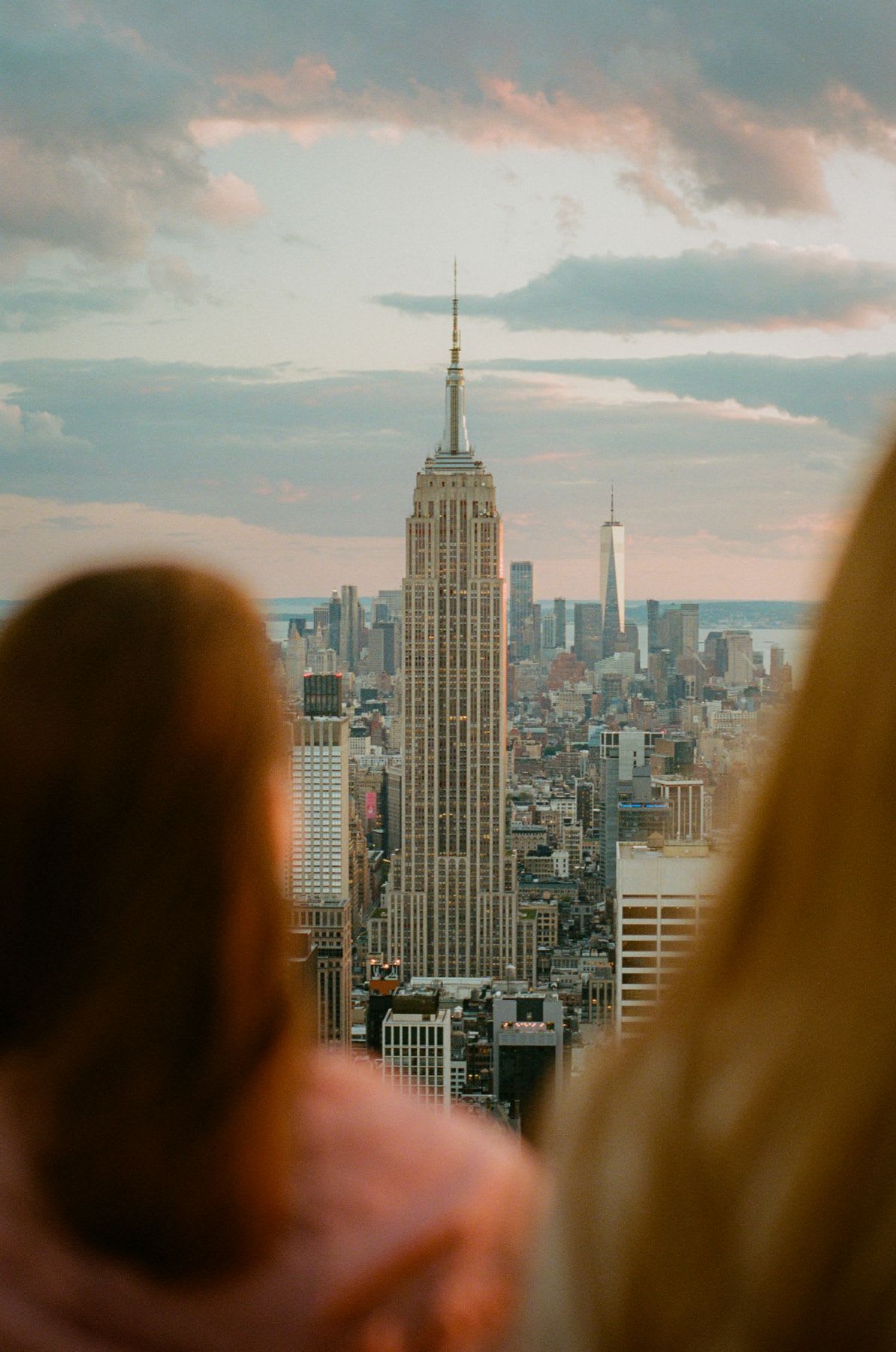 Two women standing overlooking the New York City skyline with the Empire State Building in the foreground, while the One World Trade Center rises in the background, under a colorful sky at sunset.