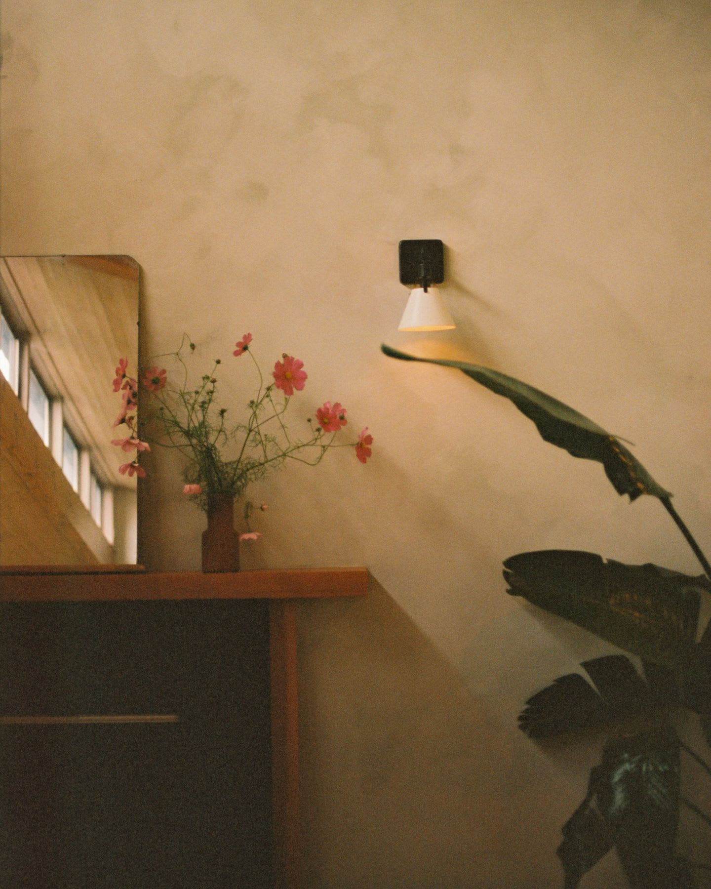 A flower arrangement next to a mirror on a shelf with a wall sconce and plant next to it.
