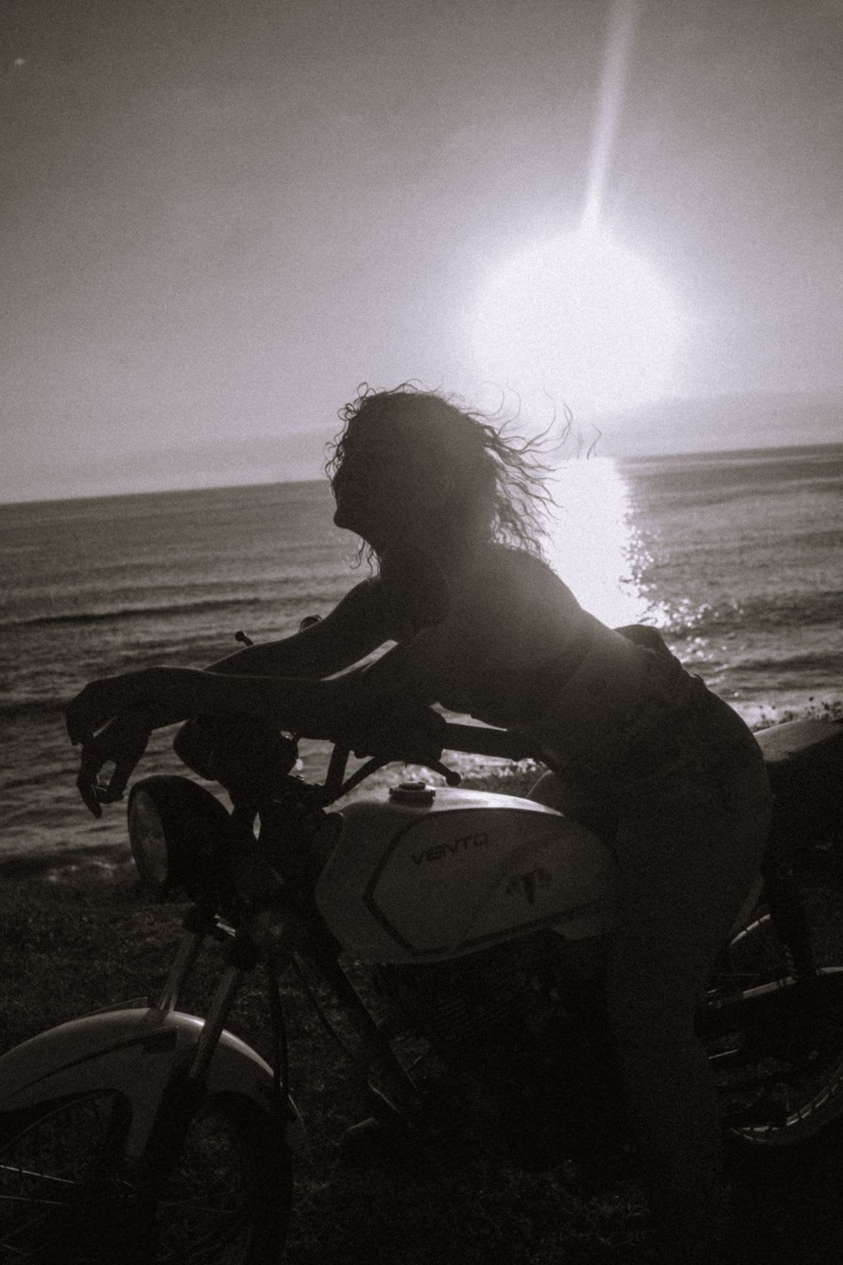 A woman with windblown hair is silhouetted on her motorcycle, enjoying a moment of freedom by the ocean against the bright sun.