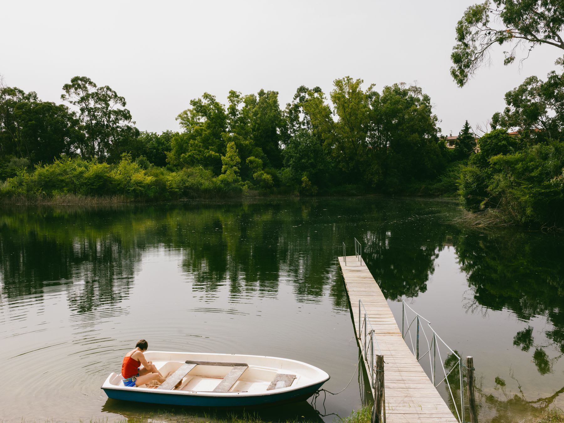 A person in a red tank top and blue shorts sits in a small white rowboat near a wooden dock on a calm lake surrounded by green trees.