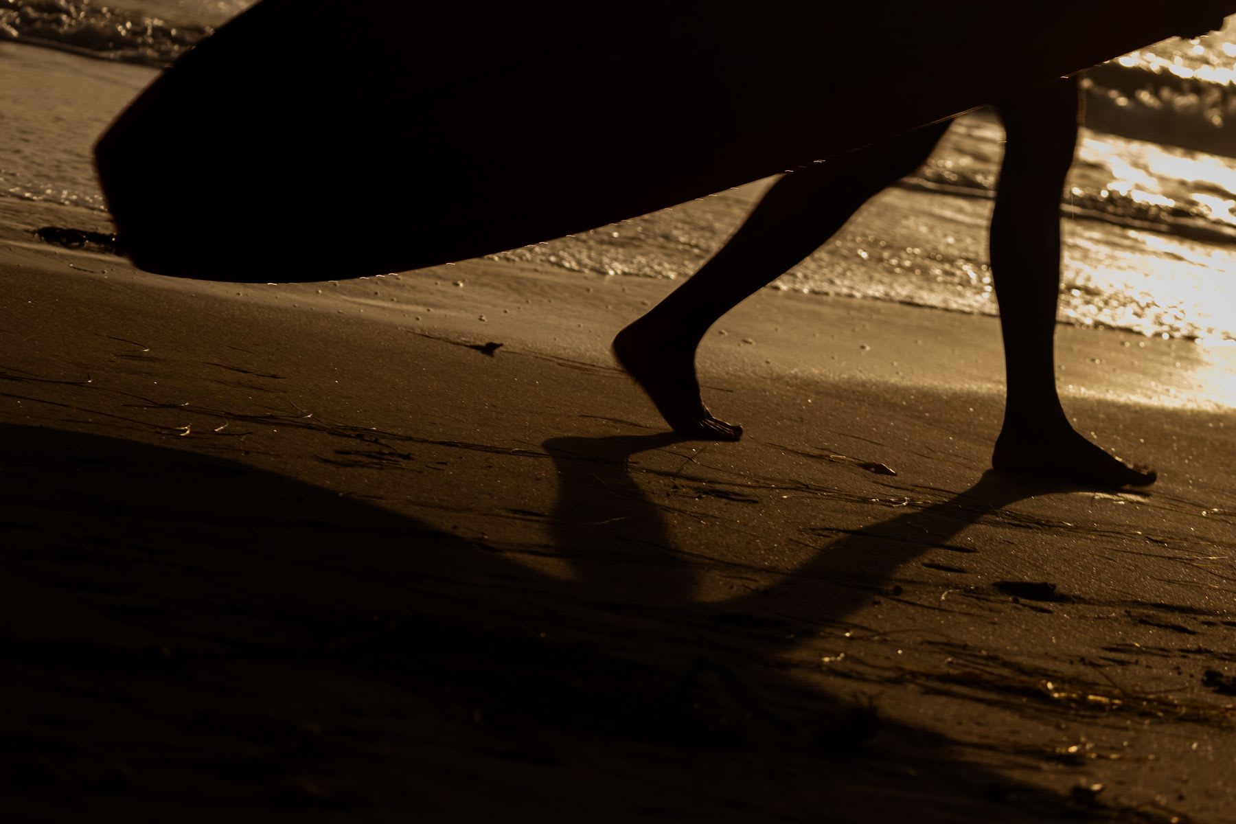 A silhouetted surfer carries a surfboard while walking barefoot on the wet sand at the shoreline during sunset.