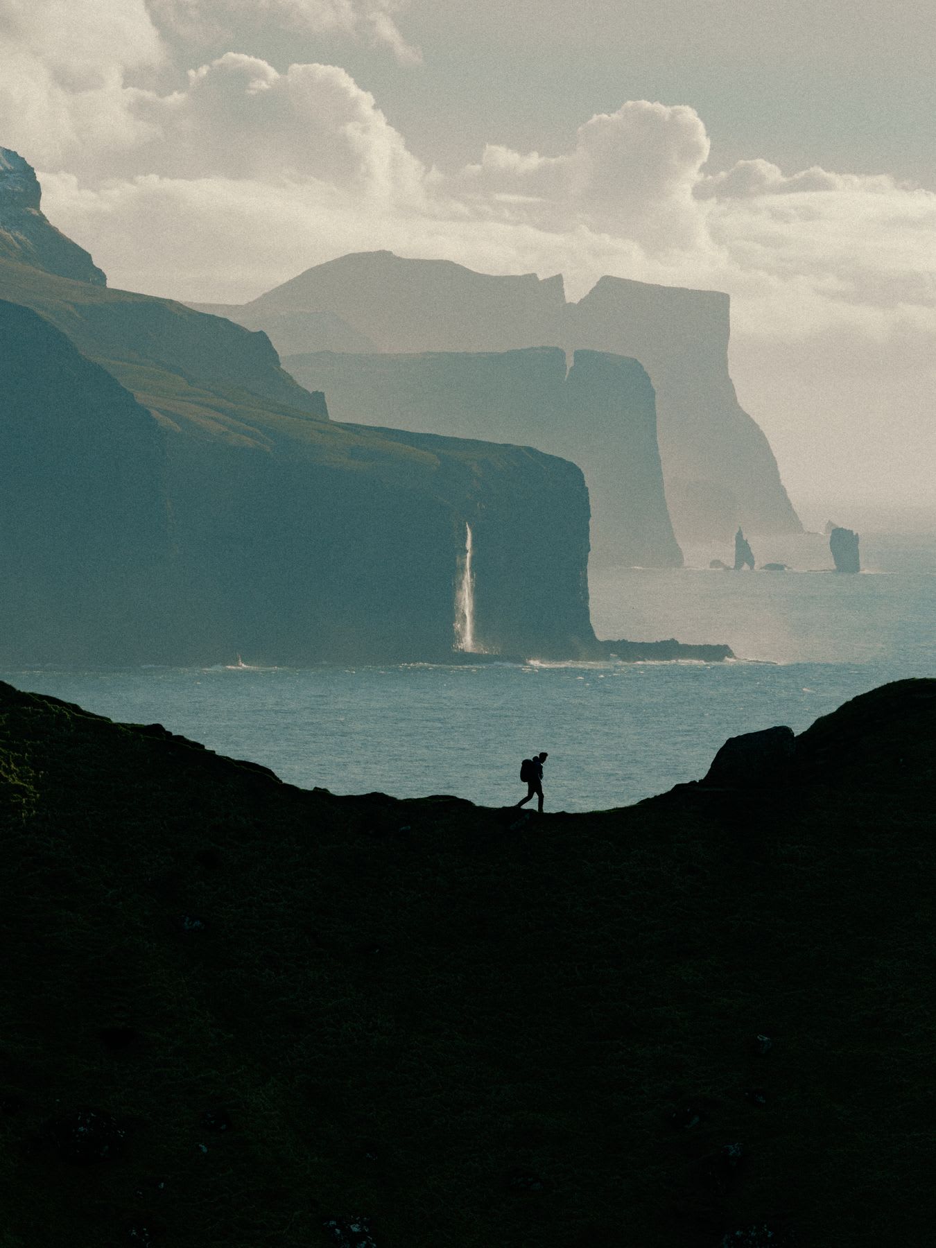 A lone hiker is silhouetted against a dramatic coastal landscape with towering sea cliffs and a waterfall plunging into the ocean.