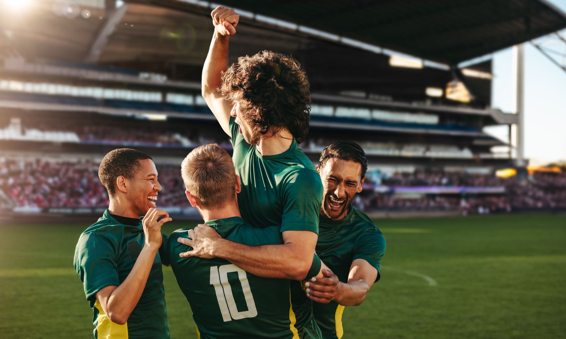 Four football players celebrating a goal during a daytime football match.