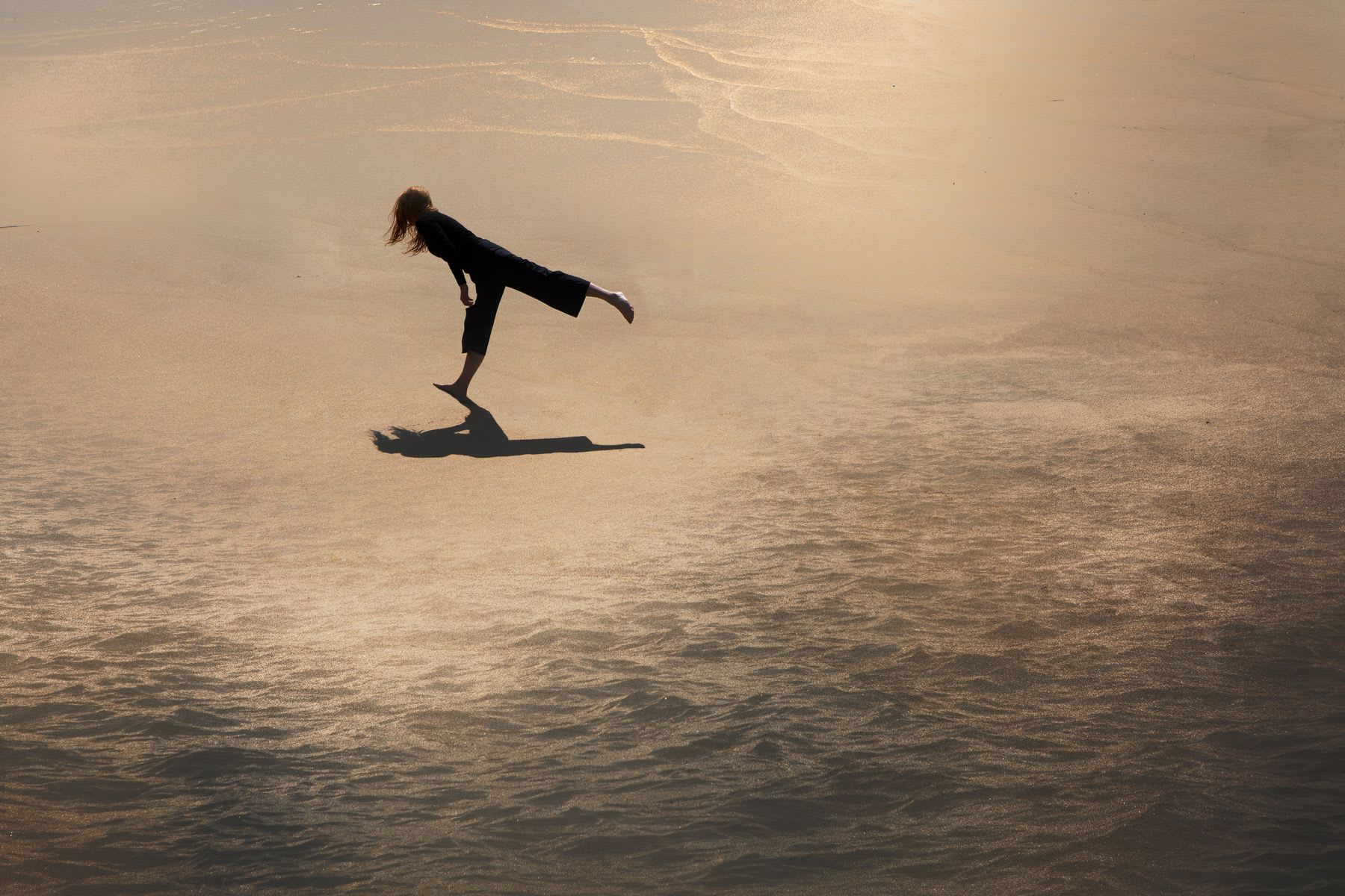 A high-angle view of a woman balancing gracefully on one leg on the glistening wet sand of a beach at sunset.