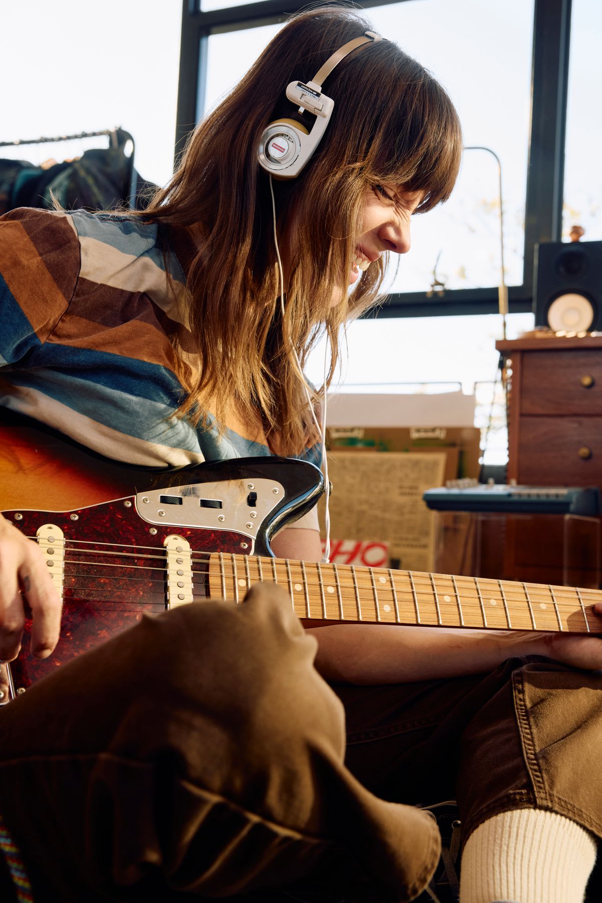 A young woman wearing vintage headphones smiles as she gets lost in the music while playing an electric guitar.