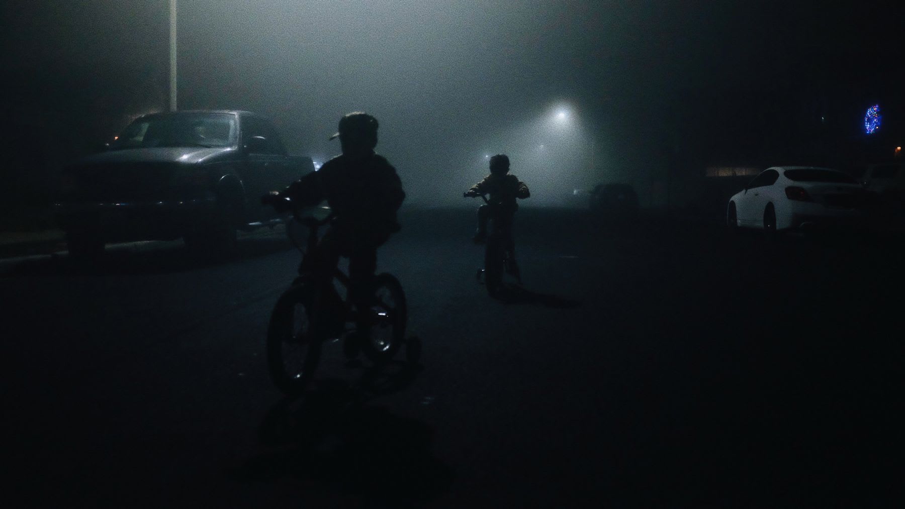 Two silhouetted children ride their bicycles down a dark street enveloped in thick fog at night.