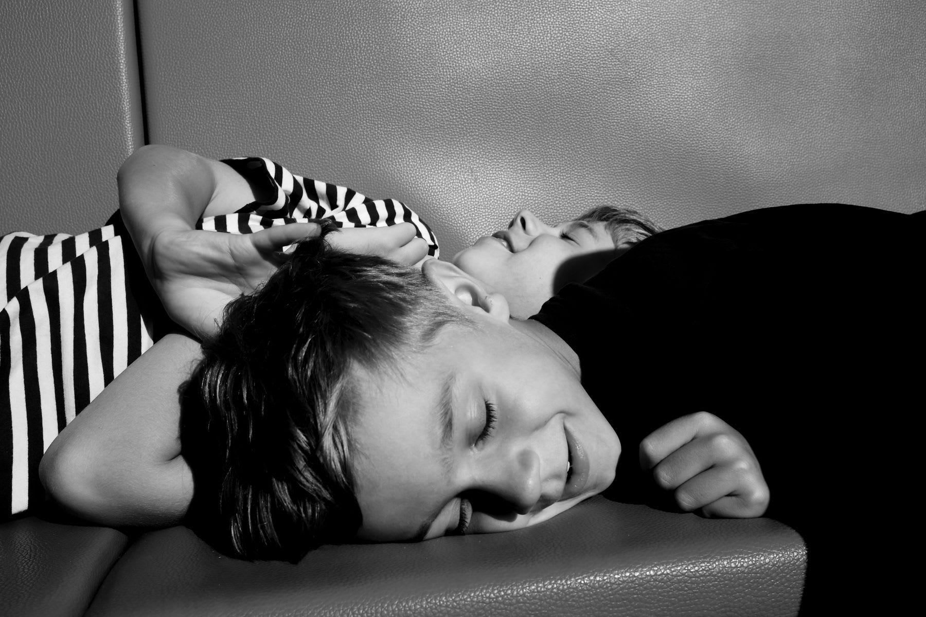 two young children lying on a cushioned surface, with one resting their head on the other’s back; both have relaxed expressions and closed eyes, suggesting happiness, warmth, and a playful bond.