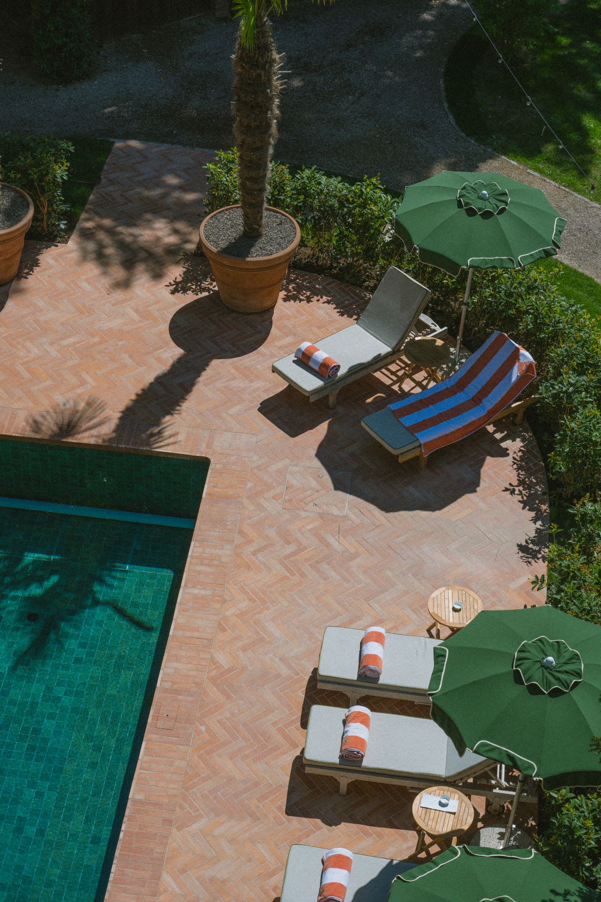 The edge of a swimming pool with several green chairs and umbrellas and a mattress made of patterned tiles