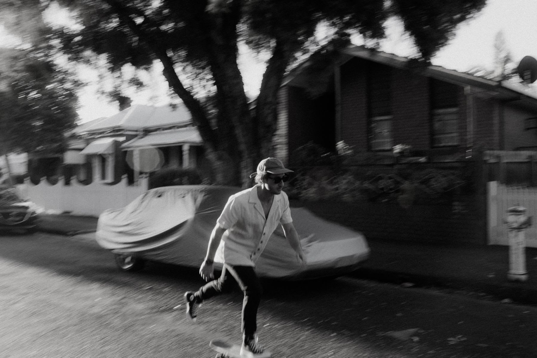 A man riding a skateboard on the street passing by a car and a house shaded by a large tree