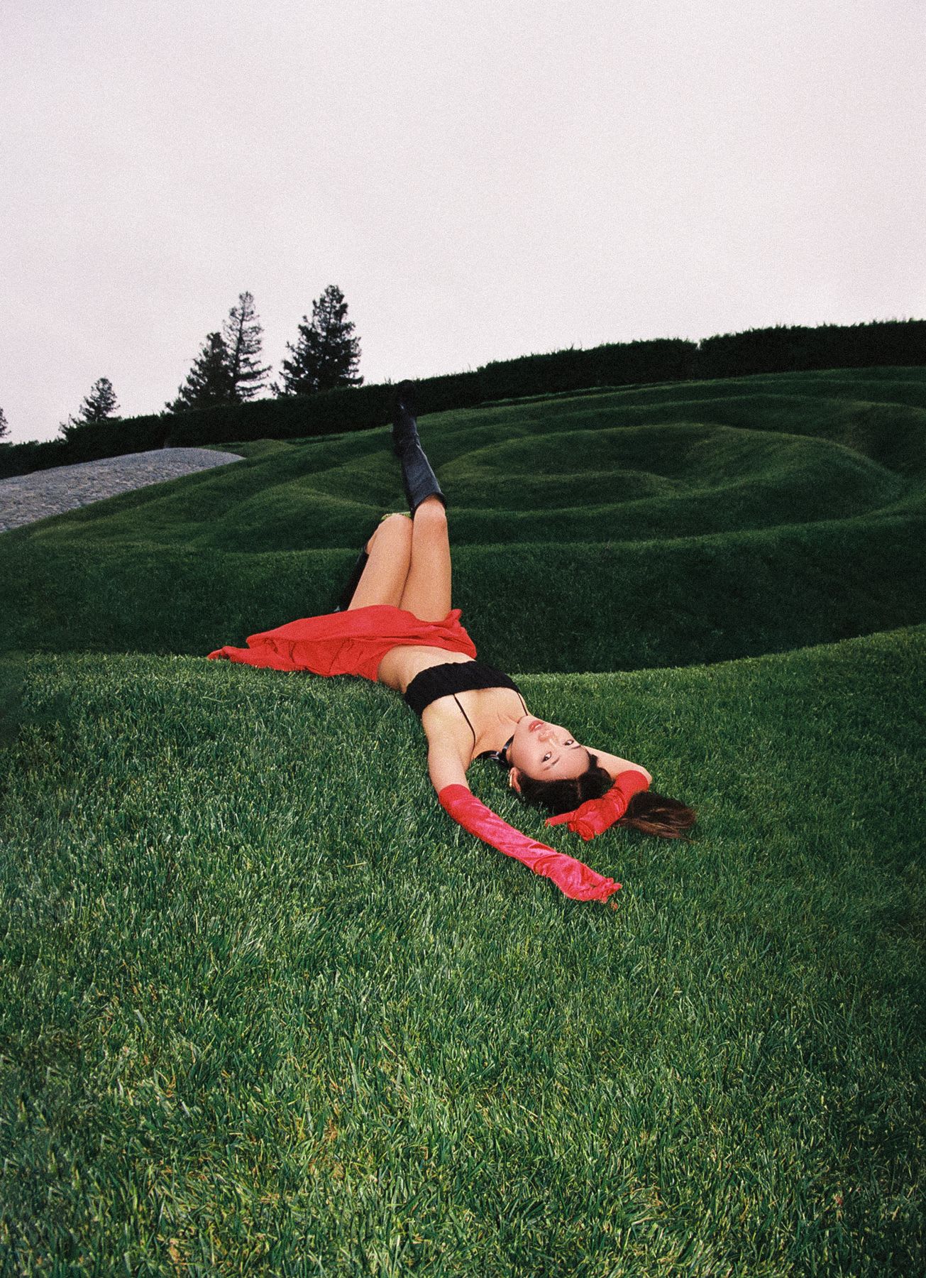 A woman in a black crop top, red skirt, and long red gloves lies on lush green grass with one leg raised, creating a dramatic pose against the backdrop of sculpted hedges and a cloudy sky.