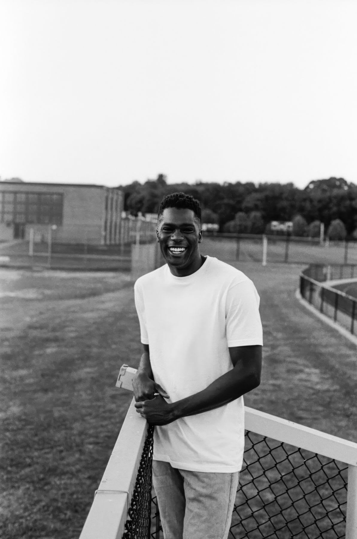 A happy young man in a white t-shirt smiles for a black and white portrait while leaning on a fence at an athletic field.