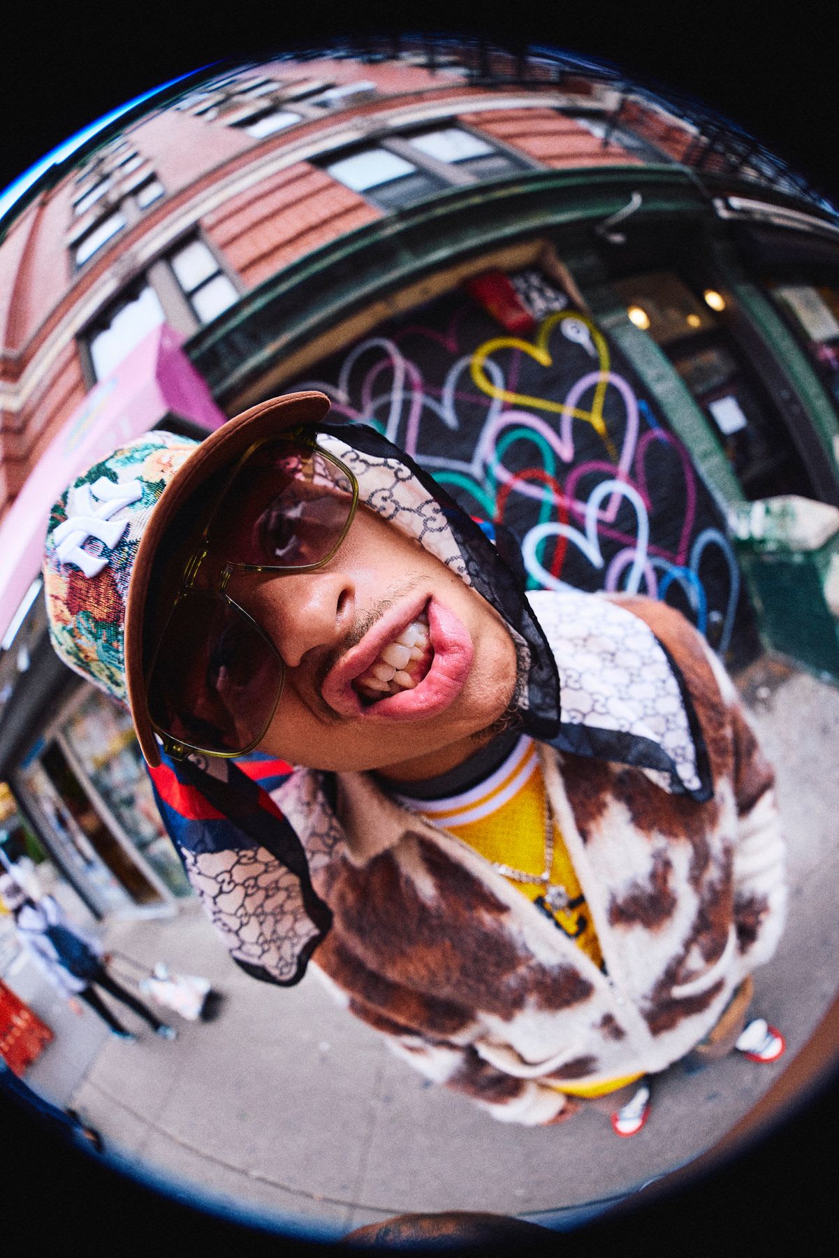 A stylish man in sunglasses and a cap smiles into the camera in a low-angle portrait taken with a fisheye lens on a city street.