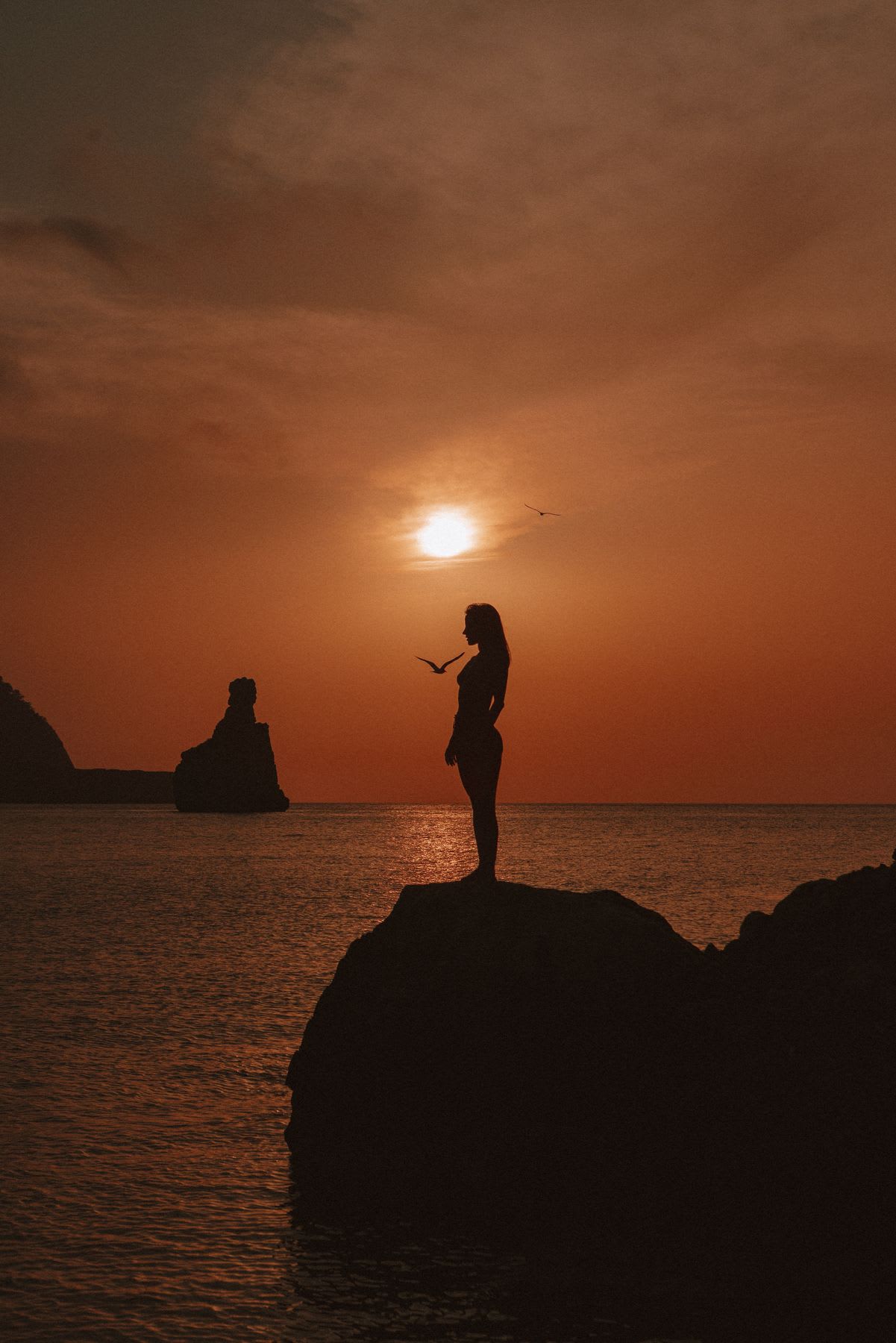 A woman in silhouette stands on a rock by the ocean, watching birds fly across the sky during a beautiful orange sunset.