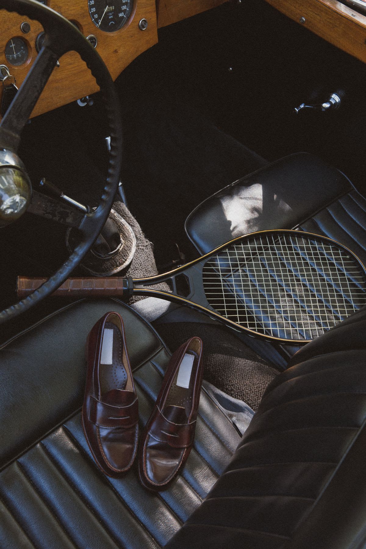 Inside a vintage convertible, a tennis racket and a pair of brown leather penny loafers sit on the ribbed black passenger seat.