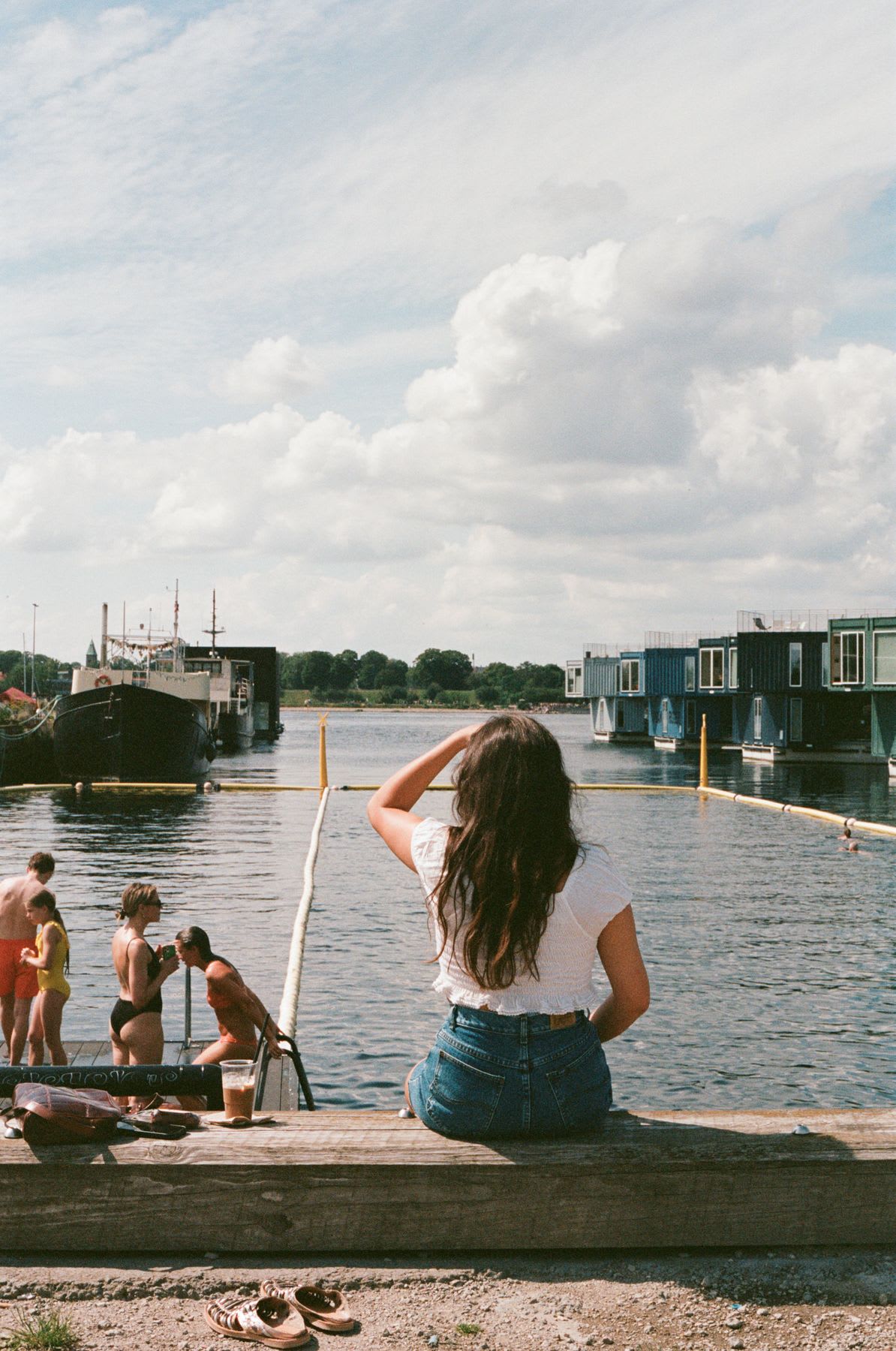 Woman sitting along the coast watching a ship in the harbor as people stand on the dock near the water