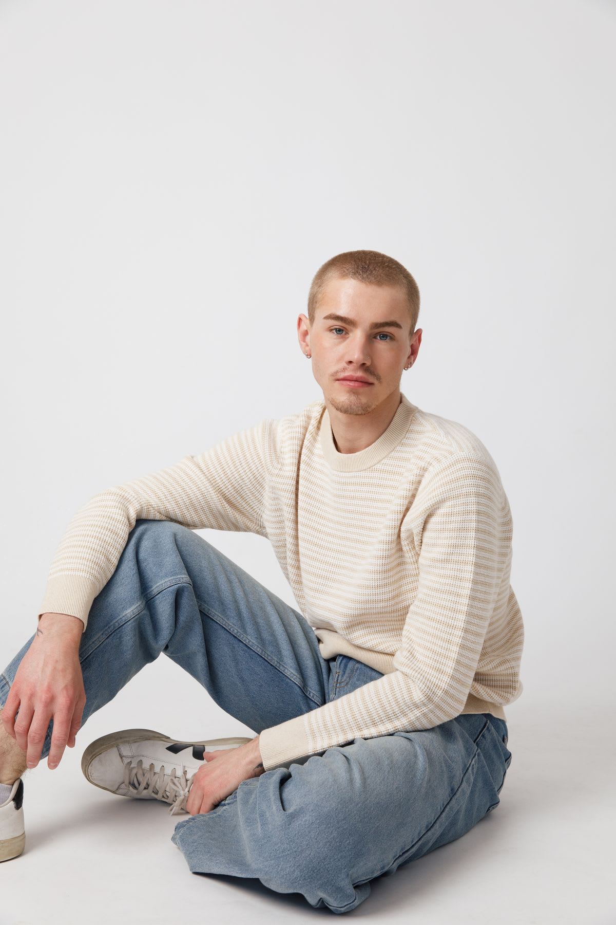A young man with a buzz cut, wearing a striped sweater and jeans, sits casually on the floor against a white background, looking directly at the camera.