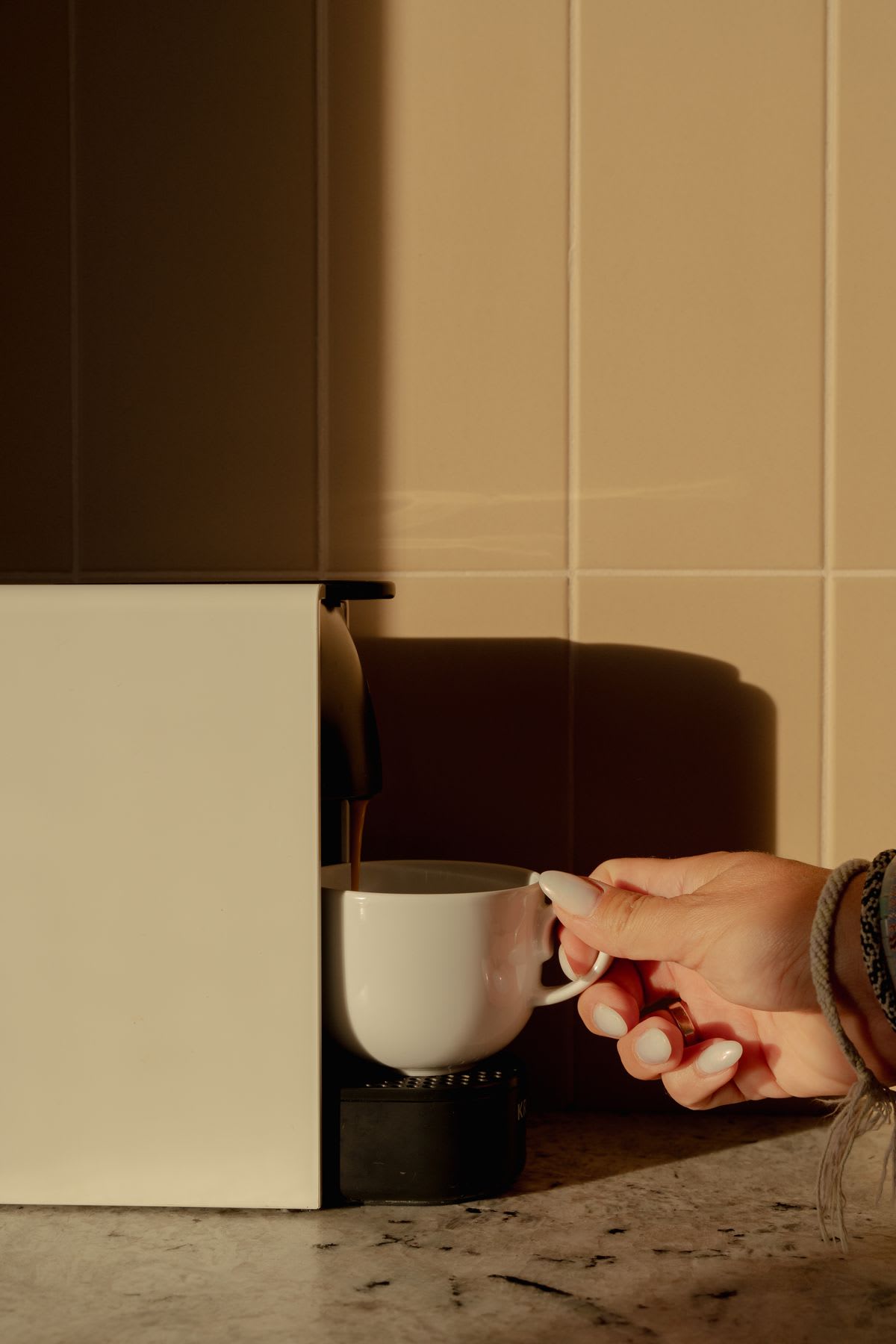 A hand holding a white cup as coffee pours from a machine, with soft sunlight casting shadows on the kitchen counter.