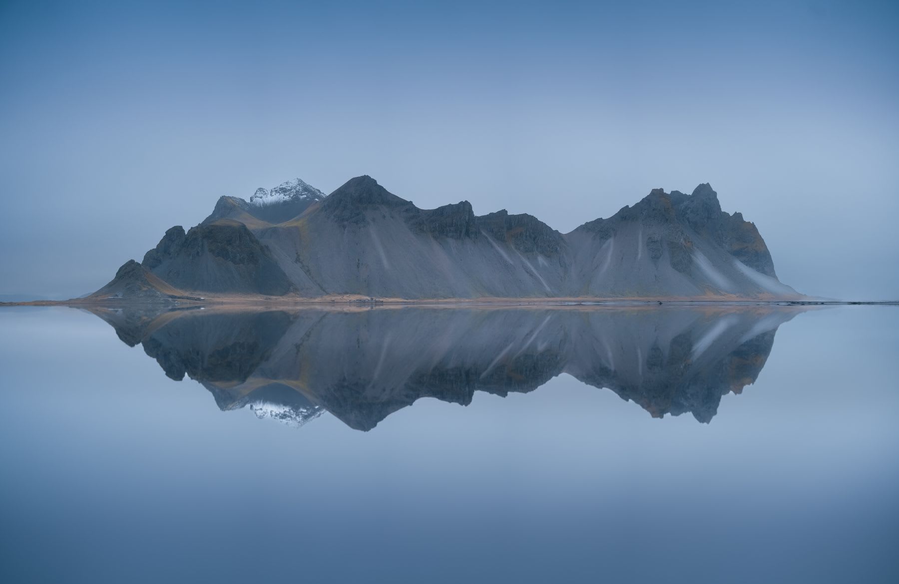A mountainous island is reflected in a still lake under foggy skies.