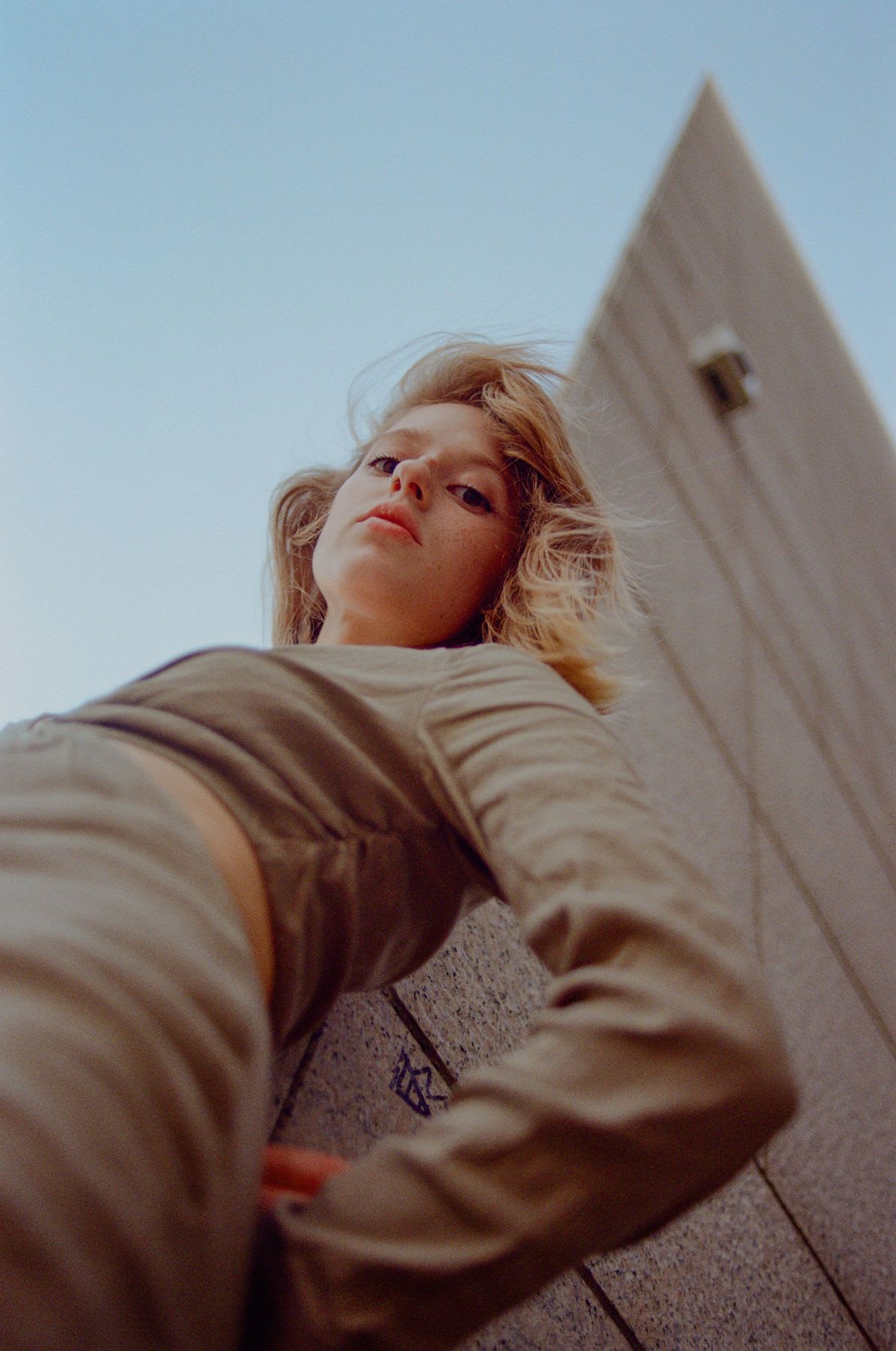 Woman poses in a fashionable outfit near the corner of a building under a blue sky.