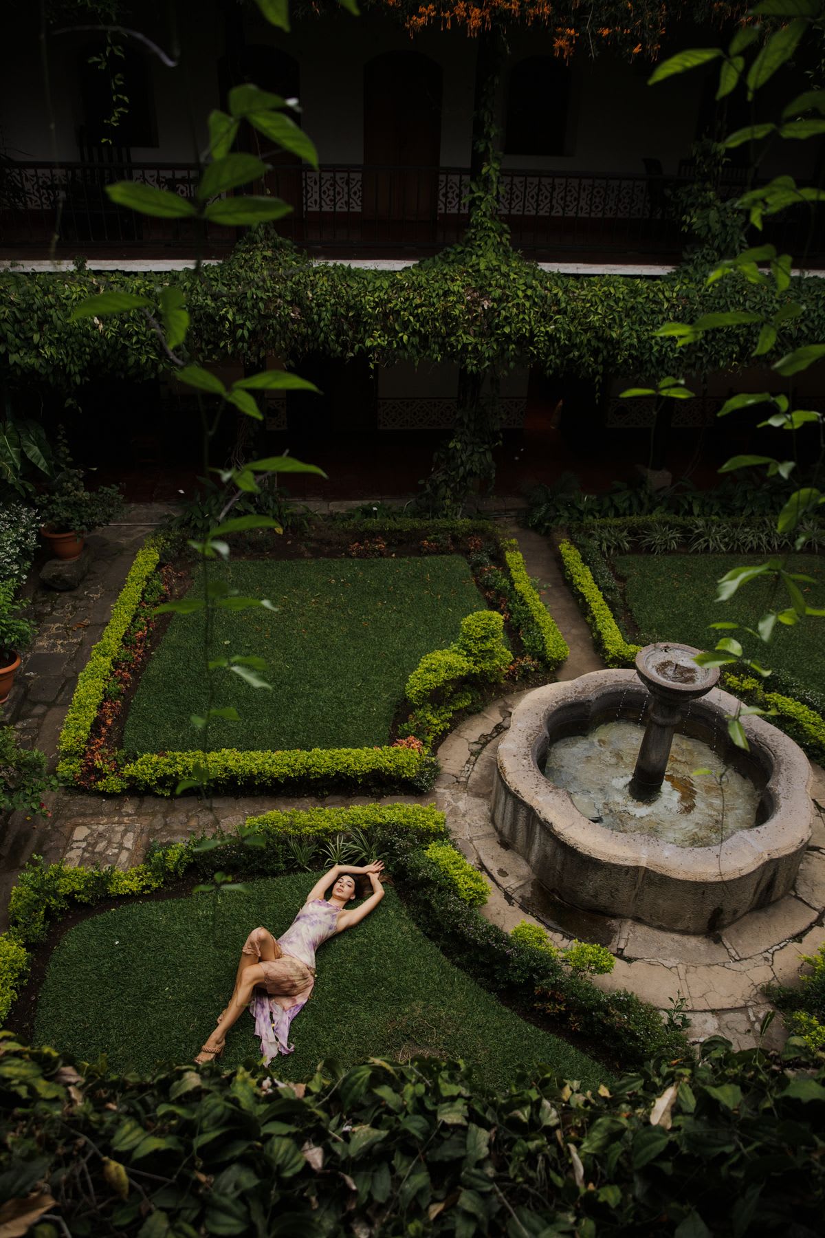From a high angle, a woman in a light-colored dress lies on a manicured lawn in a courtyard with a stone fountain, surrounded by lush green foliage and a building in the background.