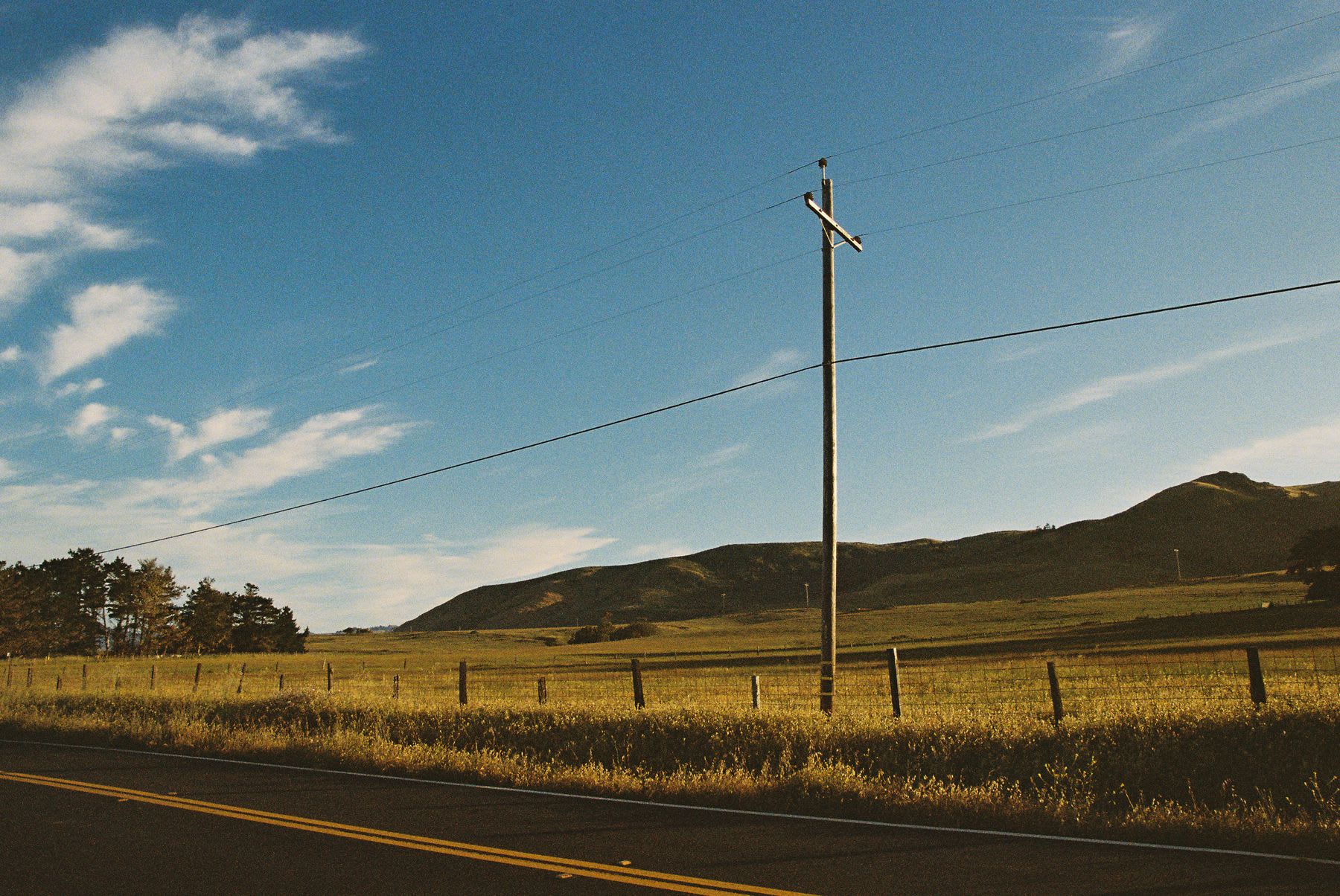 A wooden utility pole with power lines stands next to a rural road alongside a sunlit field with rolling hills in the background.