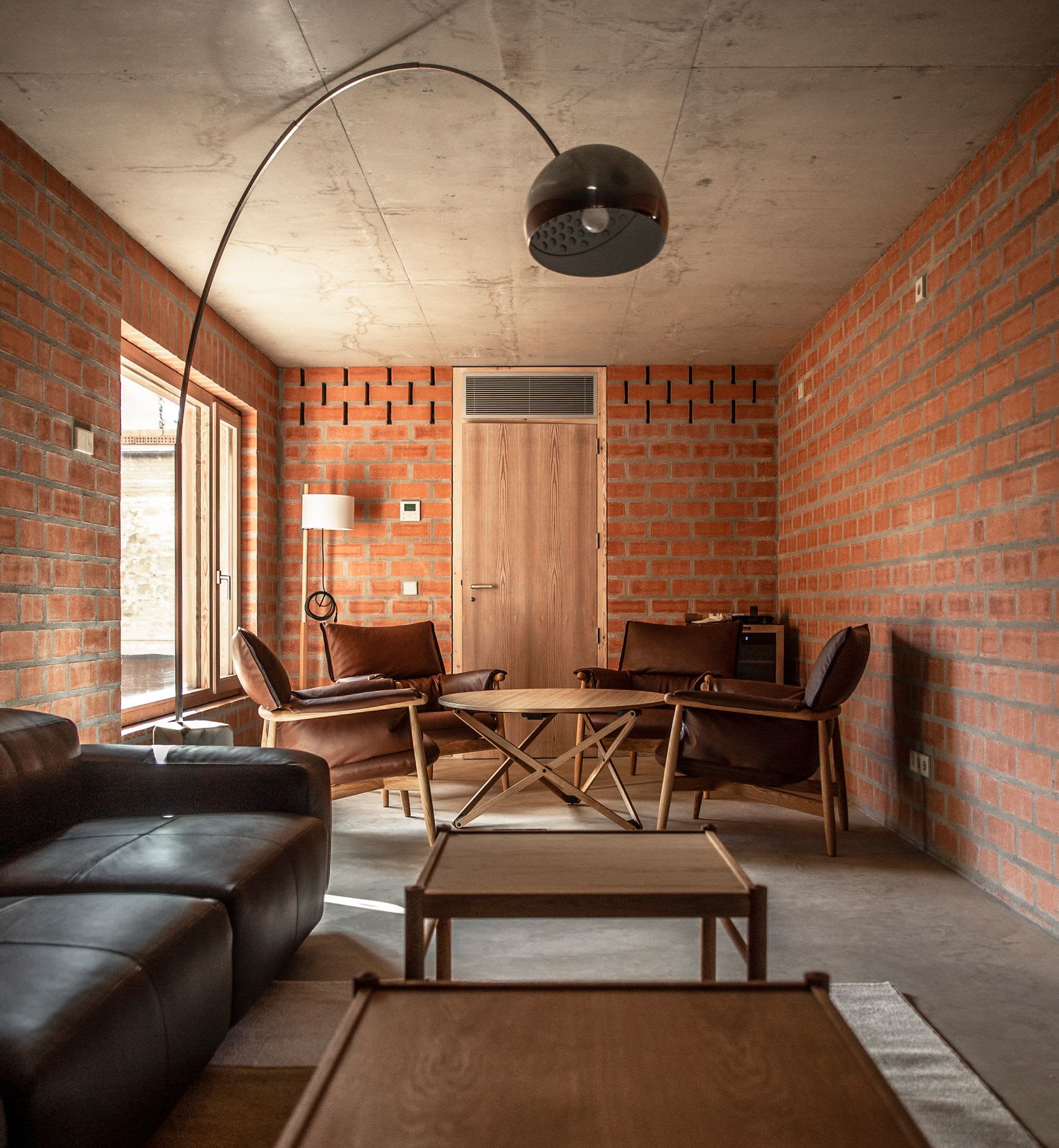 Living room with exposed brick walls and a concrete ceiling, featuring a mix of leather and wooden furniture, including a black leather sofa, brown leather chairs, and wooden tables, illuminated by natural light from large windows.