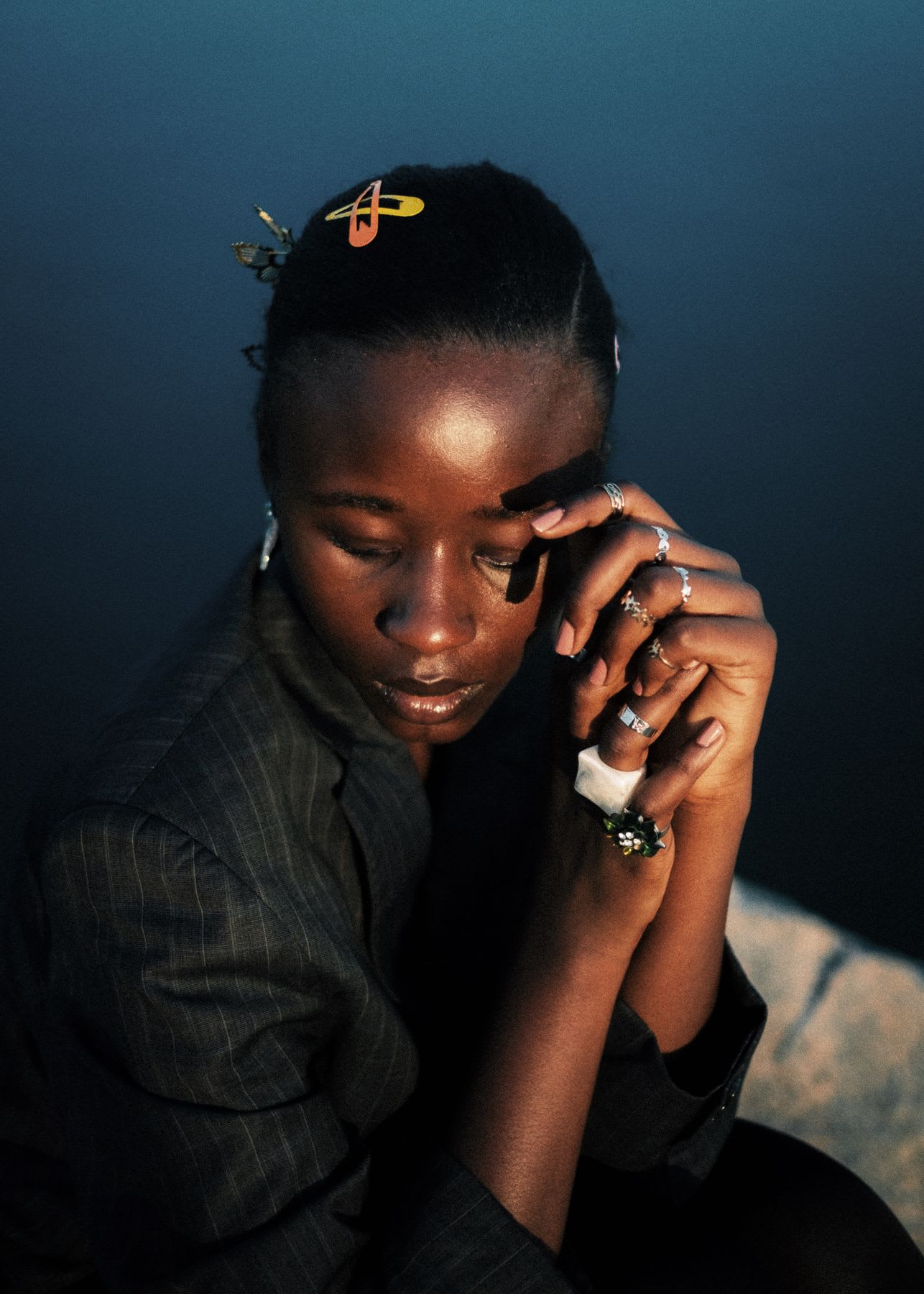 A pensive woman wearing a pinstripe blazer and multiple rings rests her hands by her face in the warm evening light.