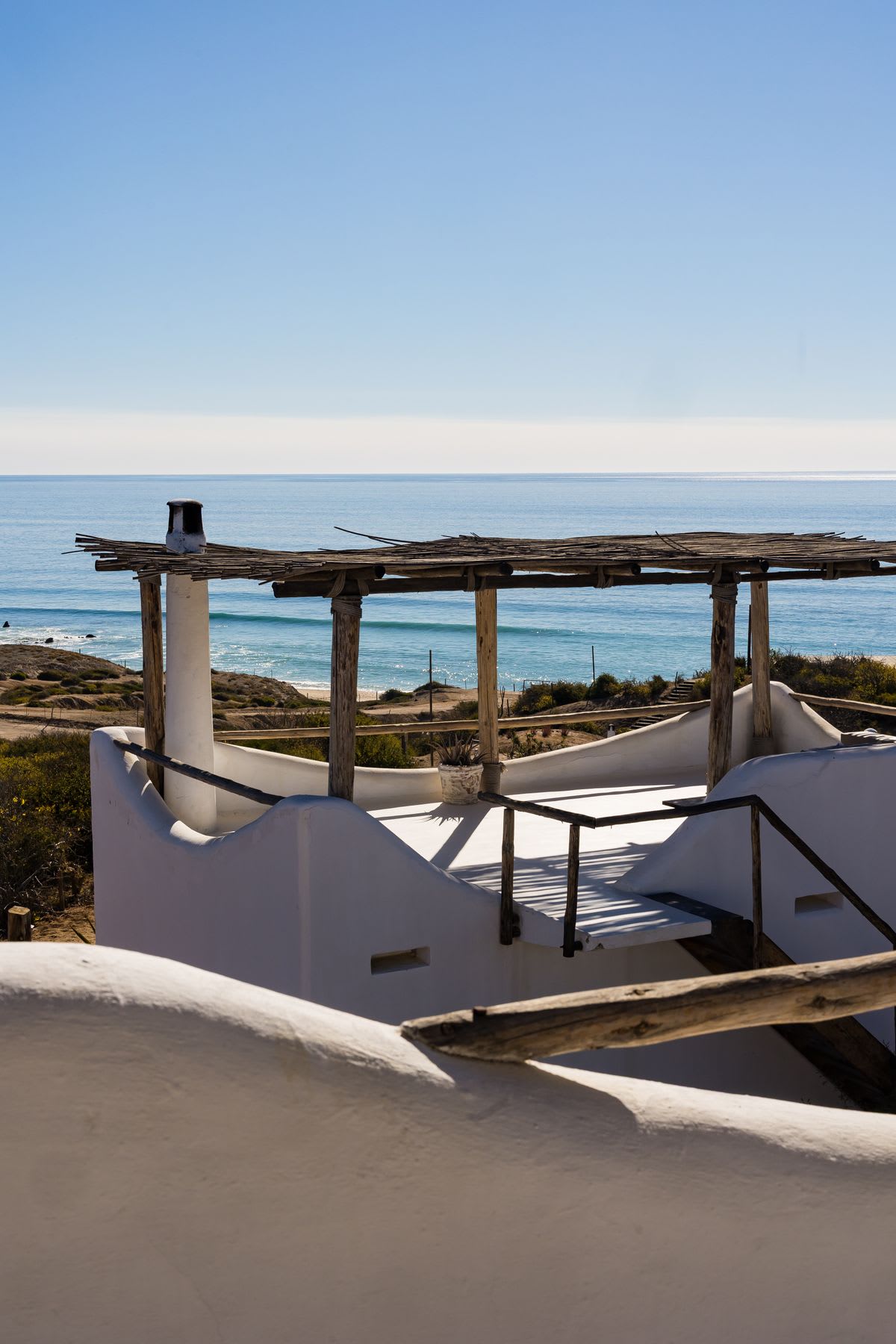 A sun-drenched white plaster building with a rustic wooden porch overlooks a beautiful blue ocean on a clear day.