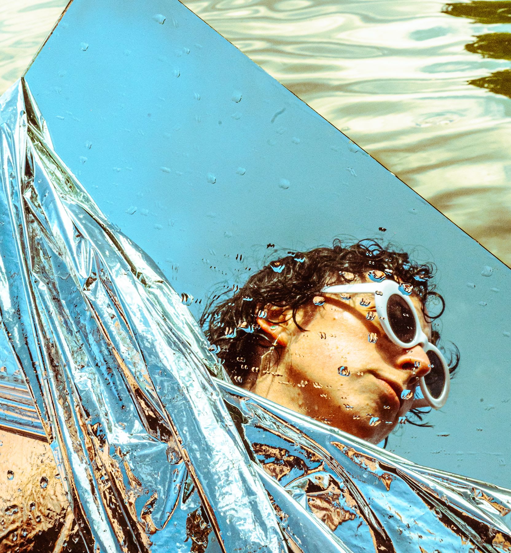 Shimmering fabric wrapped around a mirror with a person's face reflected in the glass and the surface of the water behind it all
