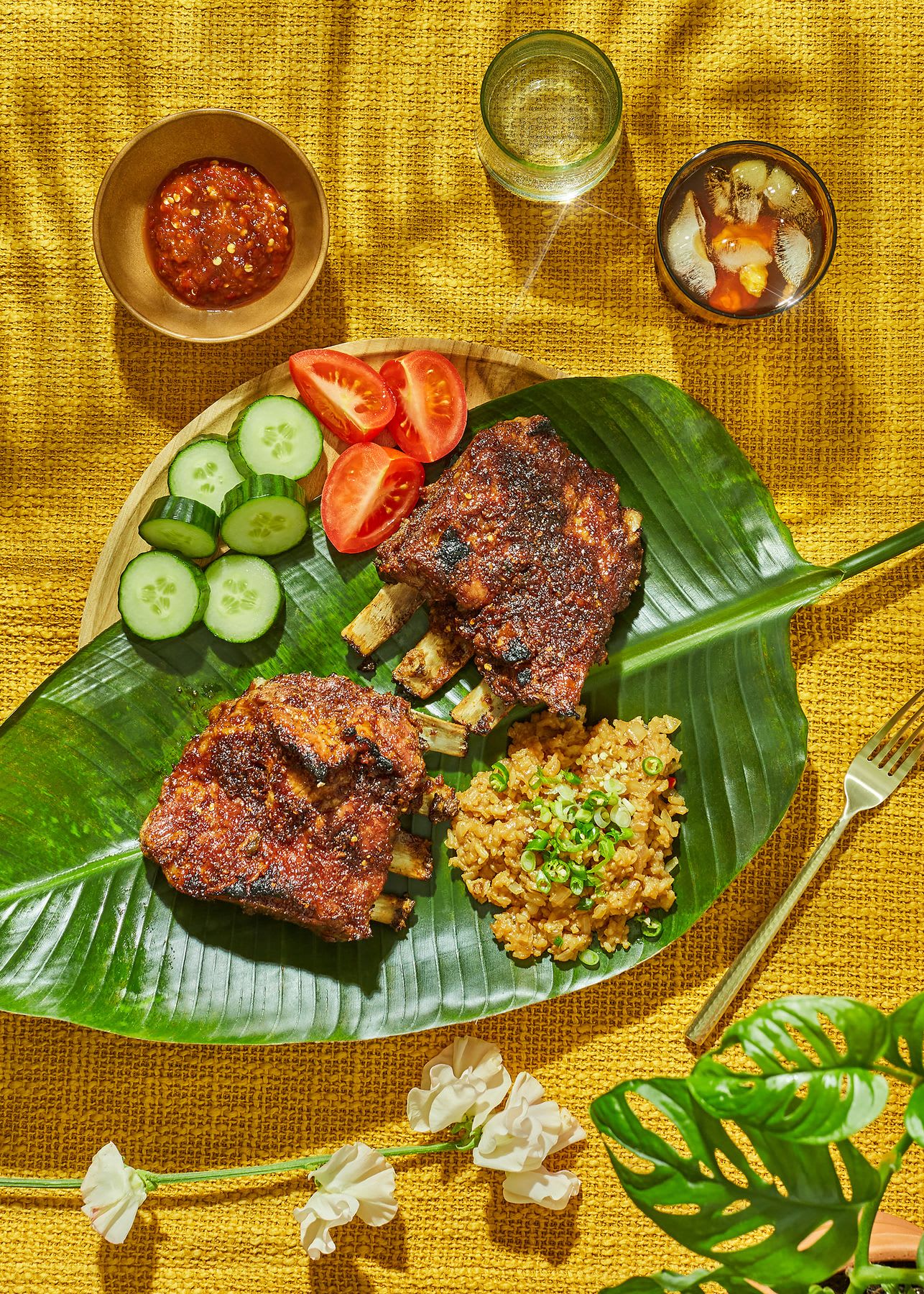 A vibrant orange tablecloth showcases a banana leaf laden with succulent ribs, fluffy rice, and colorful toppings, accompanied by a fork and water glasses, ready for a delightful feast.