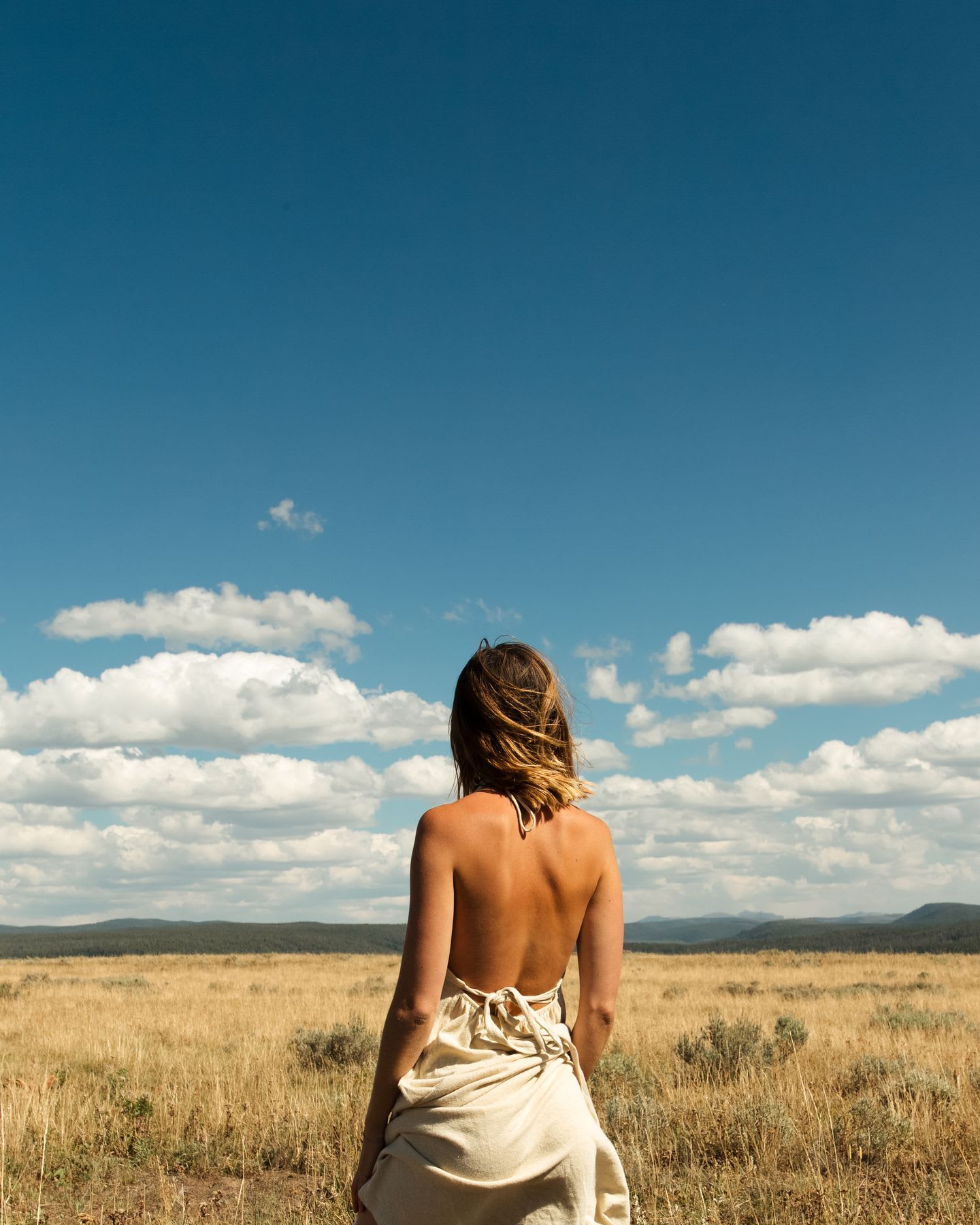 Woman wearing light summer dress stands in field bordered by mountains on the horizon.