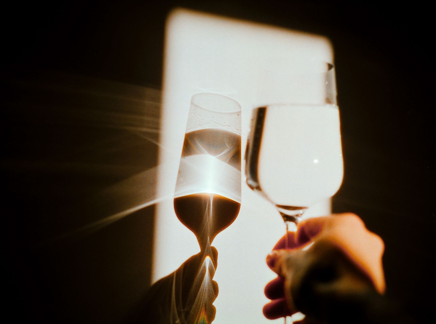 Two hands clink wine glasses together in a celebratory toast, beautifully illuminated by a stark beam of light causing a lens flare.