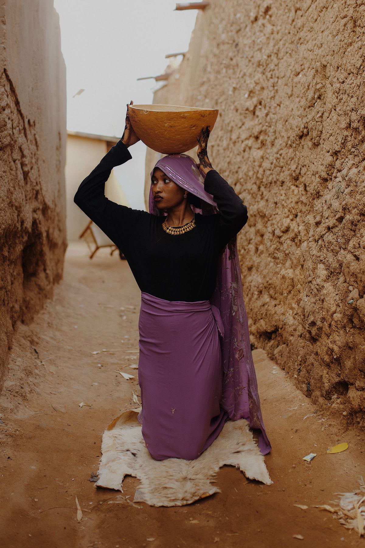A woman in a purple headscarf and skirt kneels in a narrow earthen alleyway while balancing a large bowl on her head.