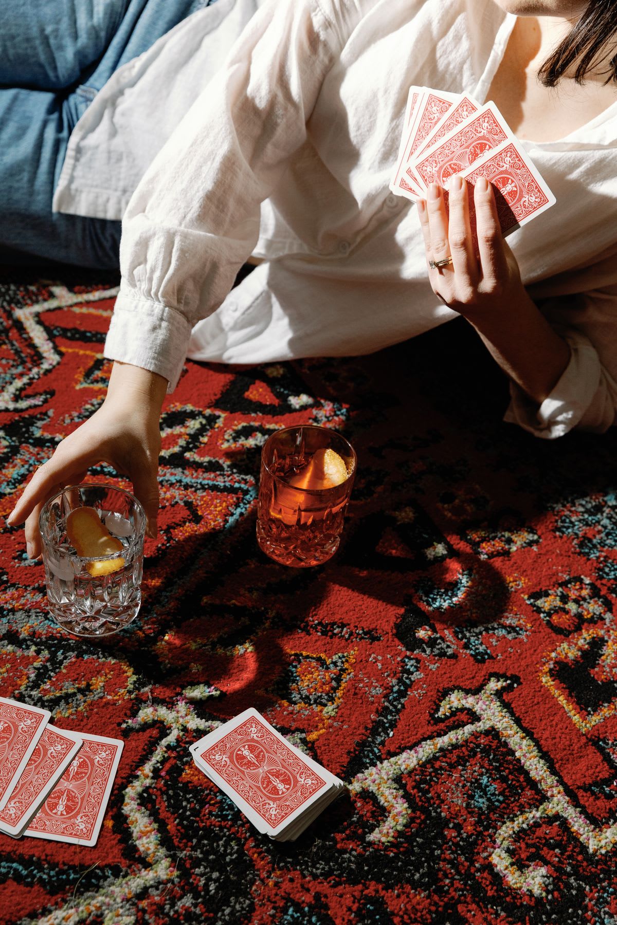 Person lying on a red patterned carpet holds playing cards in one hand while reaching for a glass of drink and ice in the other.