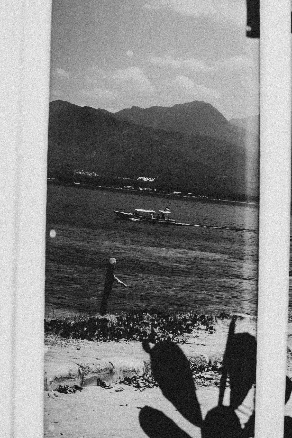 The black and white photo captures a serene coastal scene with a boat sailing past a mountainous backdrop, framed by a window with a plant casting a shadow in the foreground.