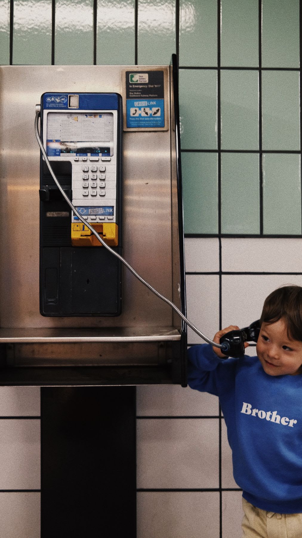 A young boy wearing a blue sweatshirt with the word "Brother" on it smiles while holding the receiver of a public payphone to his ear.