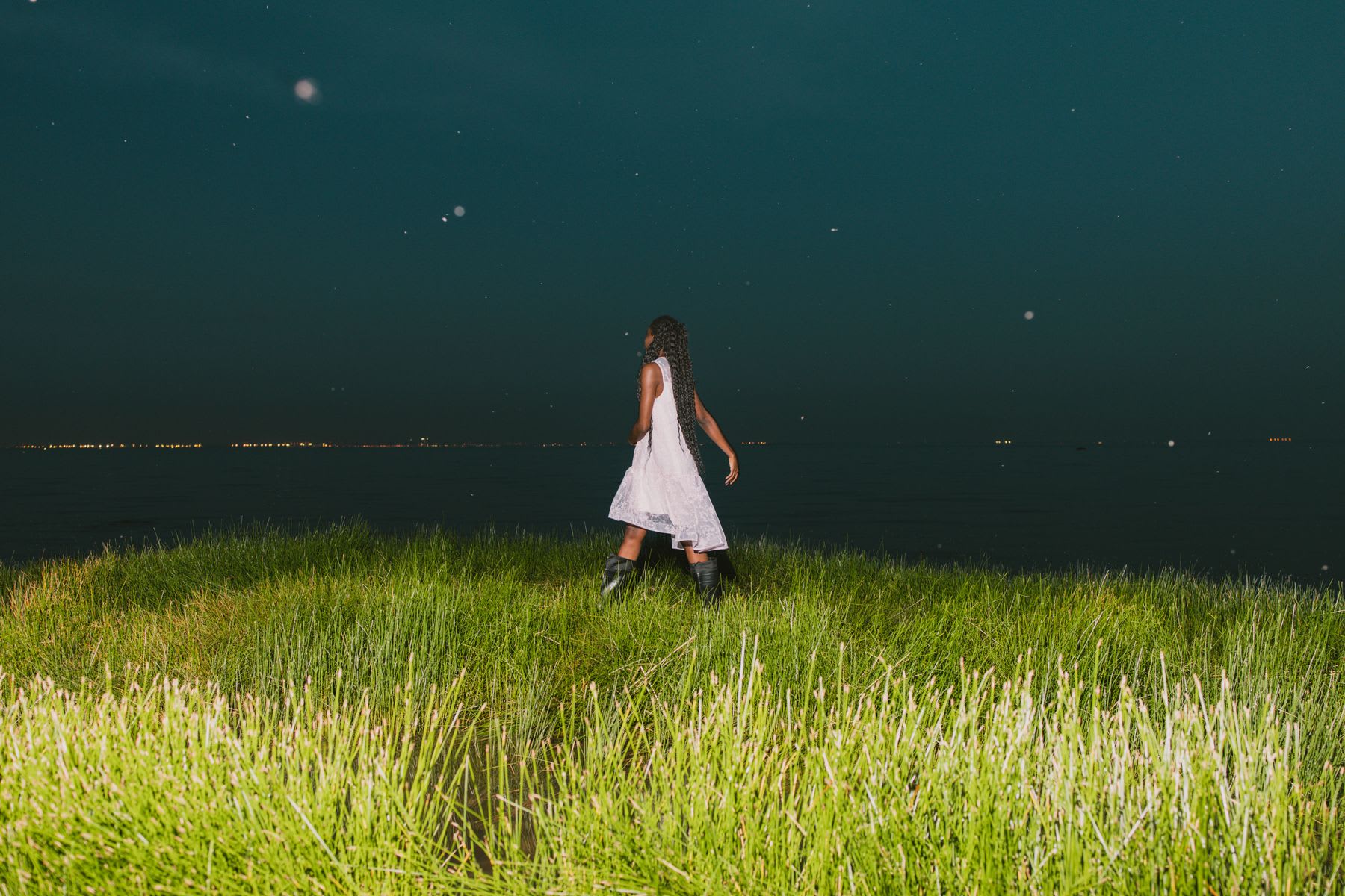 Person in a white dress and black boots walks through tall grass at night, with the dark sea and distant lights on the horizon.