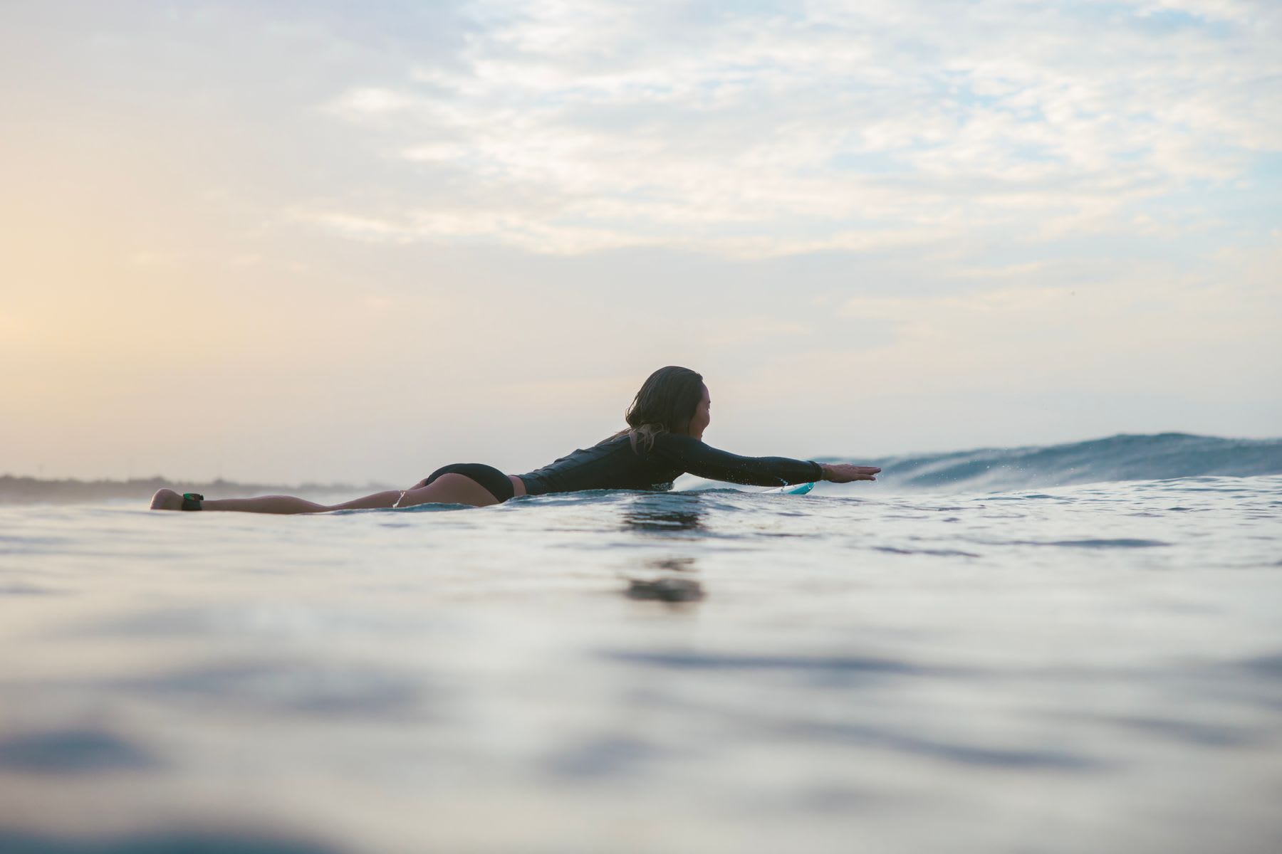 A female surfer in a wetsuit paddles on her surfboard in the ocean, waiting for a wave at sunset.