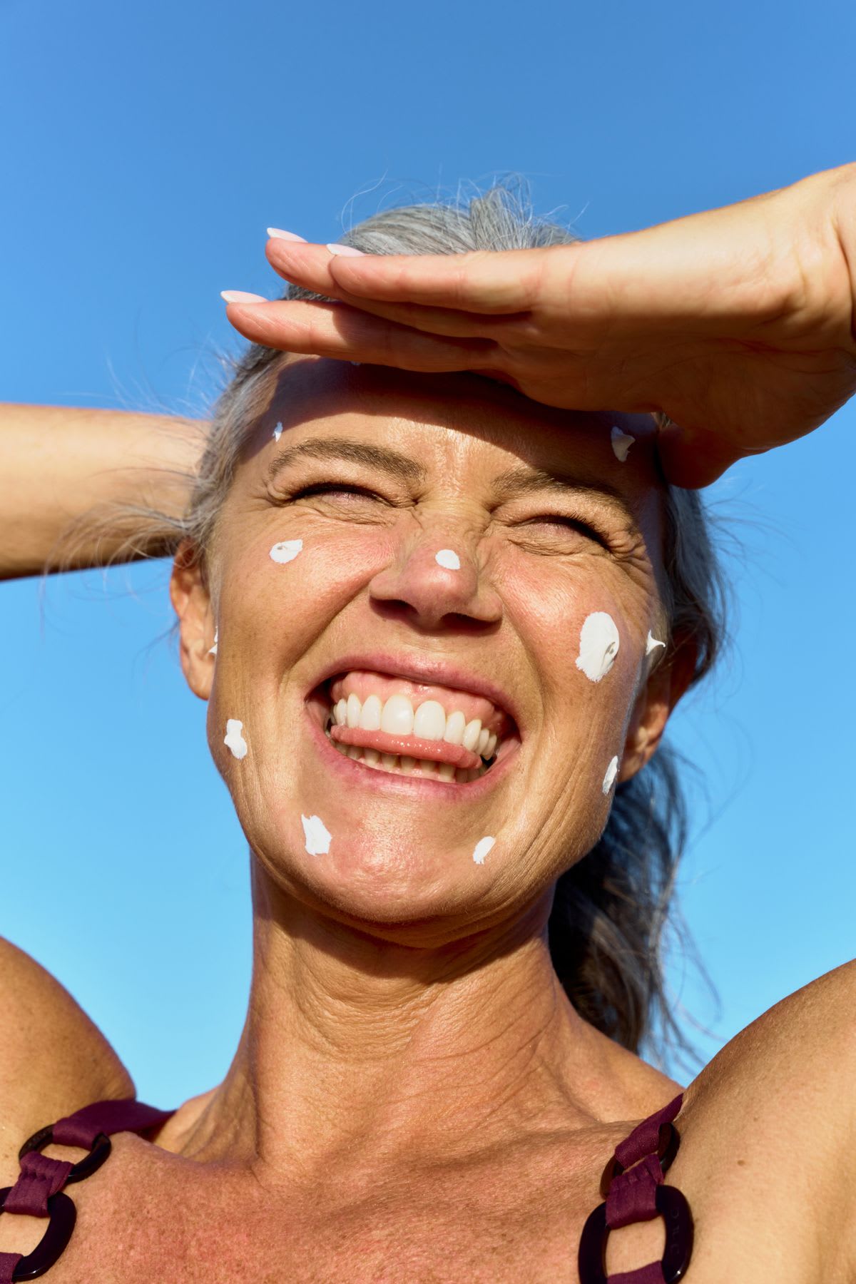 A happy mature woman with sunscreen on her cheeks shields her eyes while squinting and smiling under a clear blue sky.