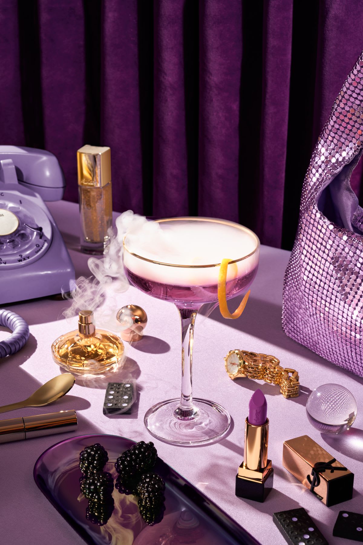 Purple-themed vanity with cocktail glass emitting smoke, surrounded by fashion accessories and vintage phone.