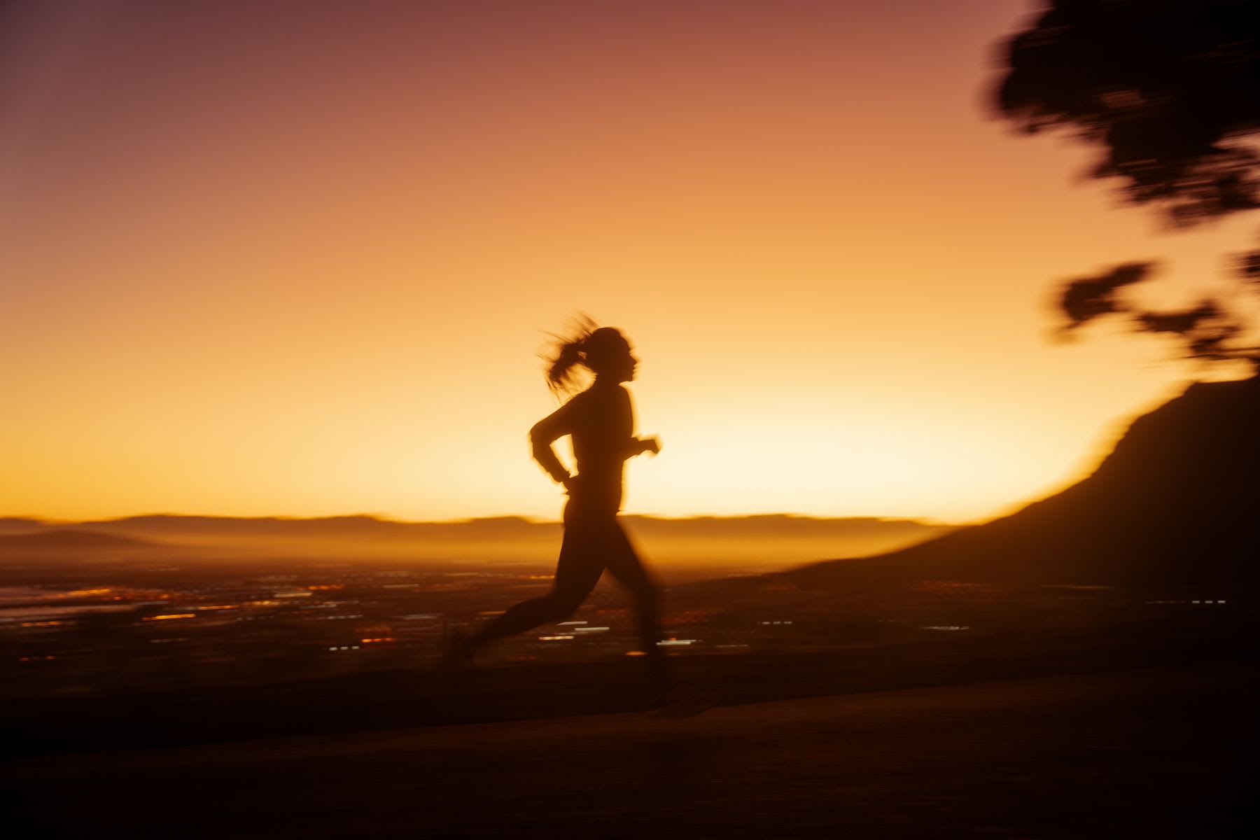A woman jogs on a mountain trail above a city at sunset.