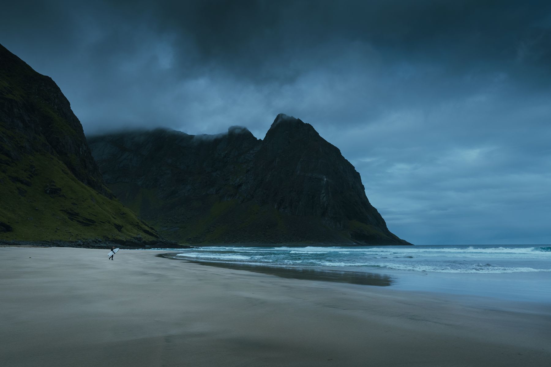 Deserted beach with smooth sand and a cloudy sky where a single person walks near the water with a surfboard.