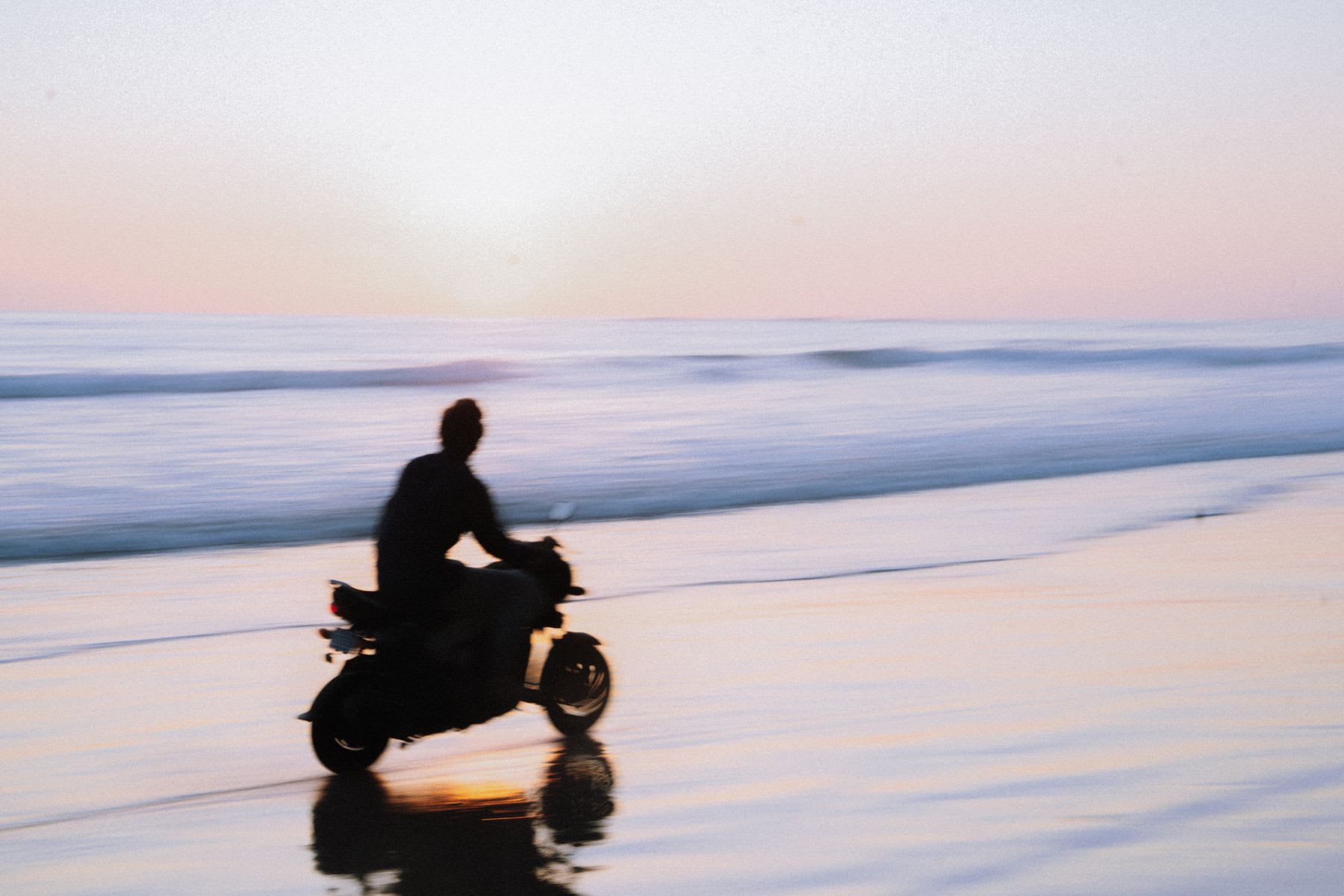 A silhouetted person rides a motor scooter on the wet sand of a beach with motion blur against the ocean at sunset.