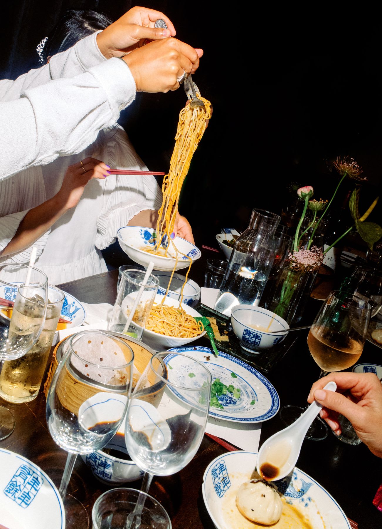 A person's hand using cutlery to pick up noodles and place them in a white bowl on a table full of utensils and glasses