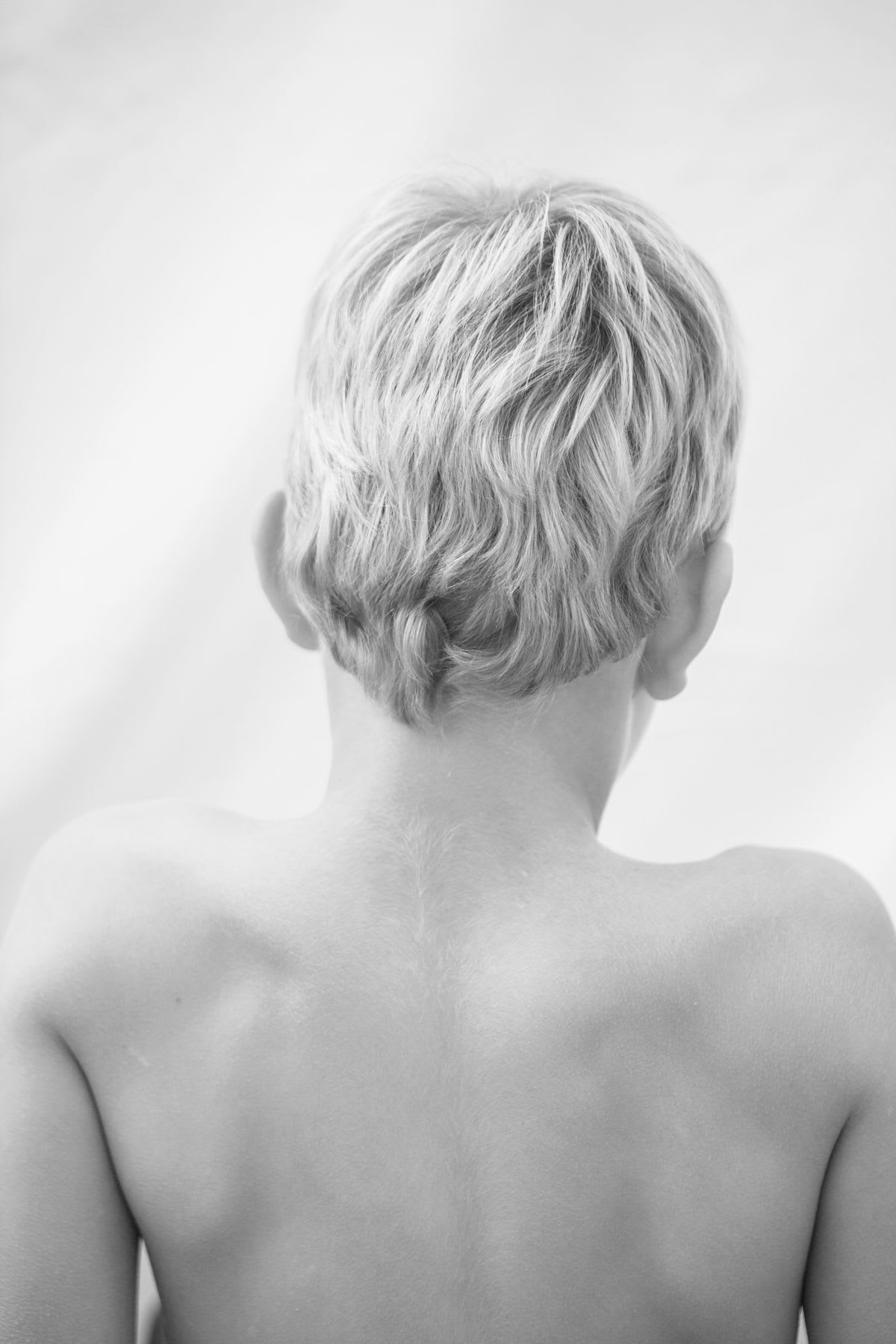 A black and white photograph captures the back of a young person with short, blonde hair and bare shoulders.