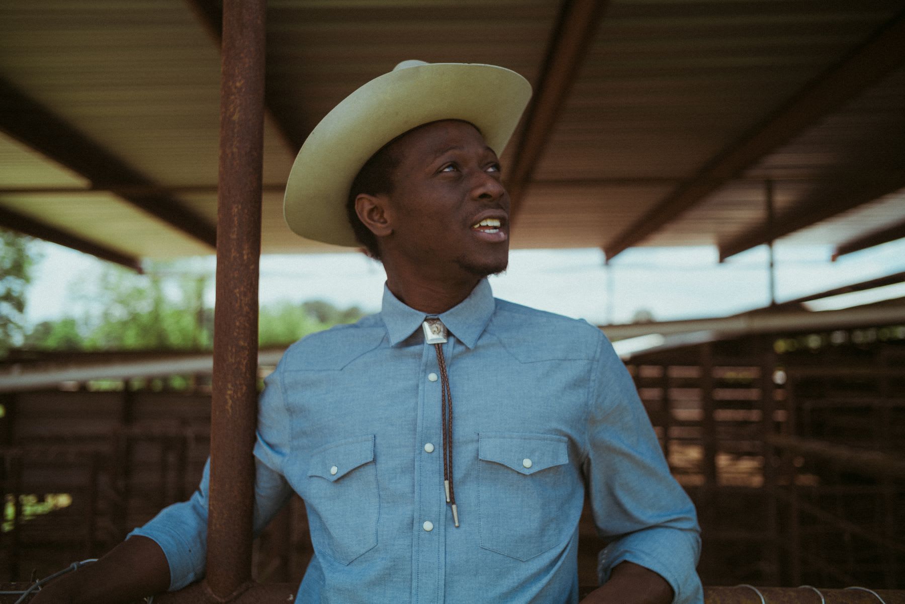 A young African American man wearing a cowboy hat and a blue western shirt with a bolo tie looks up while leaning on a rusty pole in a stable.