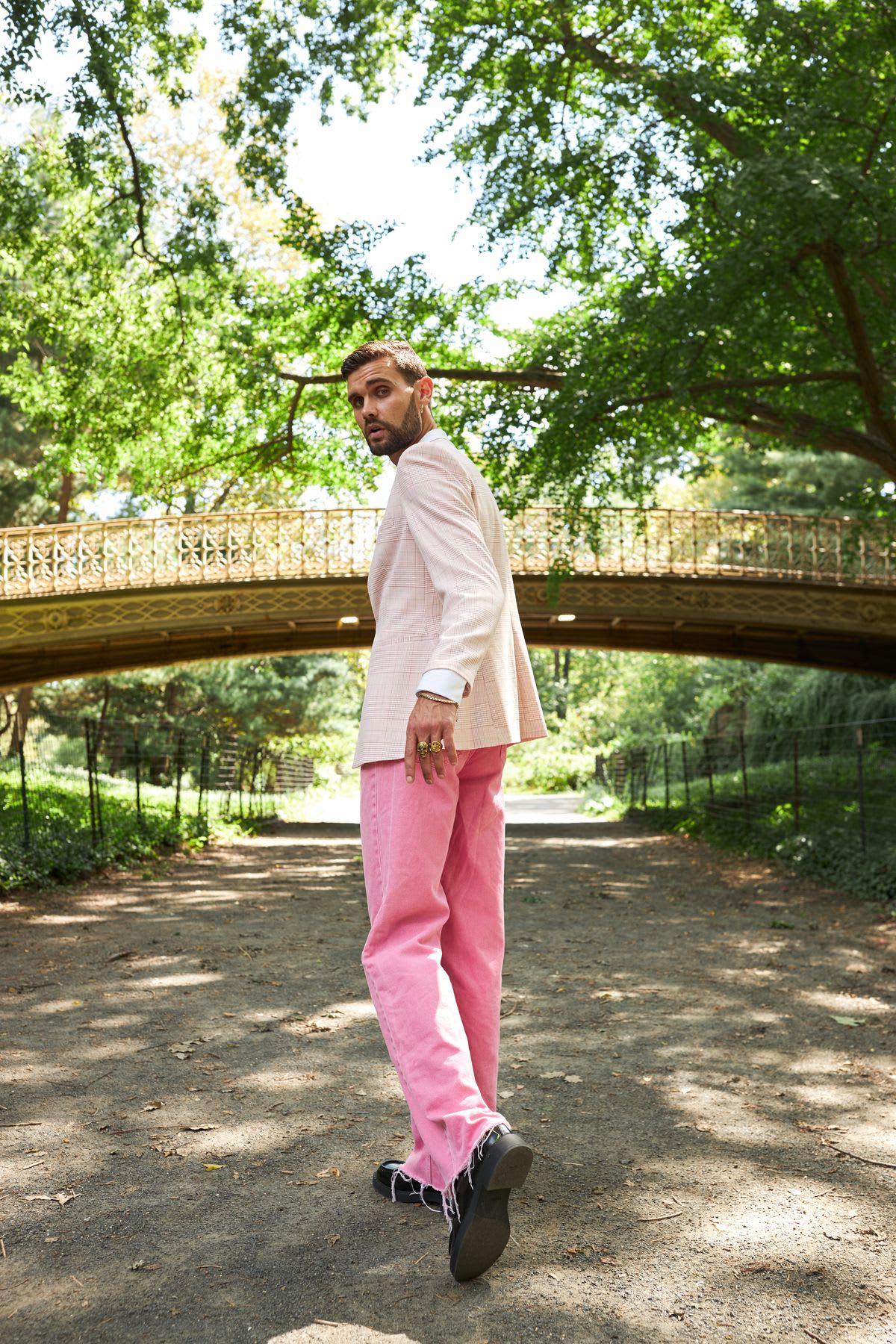 A man modeling pink jeans and a light colored jacket looks over his shoulder while walking along a park pathway.